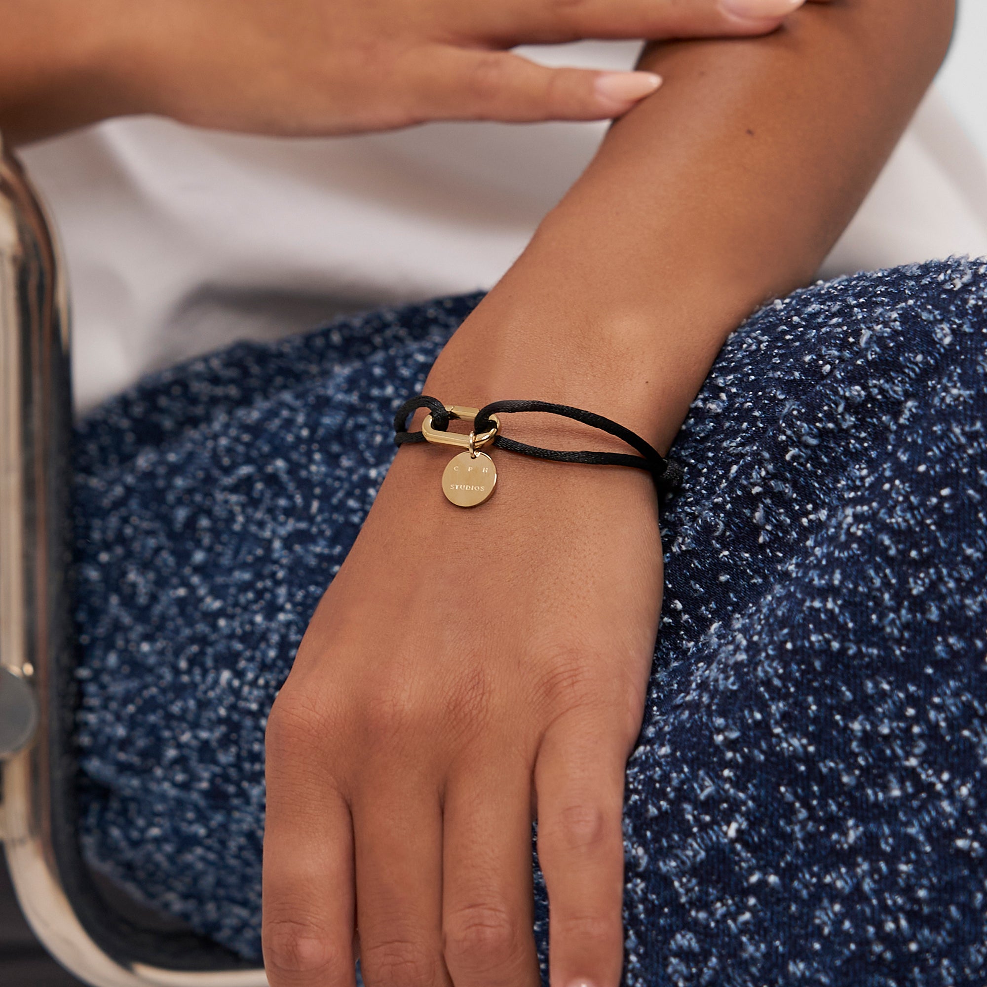 Eine Person, die ein Copenhagen Studios Armband - Fabric Link in Gold mit einer schwarzen Kordel trägt, stützt ihre Hand auf eine blaue strukturierte Hose.