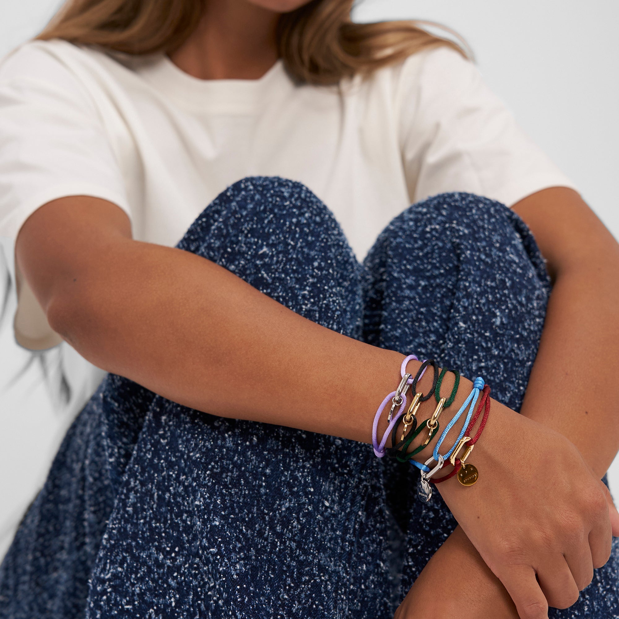 Frau in weißem T-Shirt und blauer Hose trägt mehrere bunte Schnurarmbänder, darunter das COPENHAGEN STUDIOS Armband - Fabric Link von Copenhagen Studios.