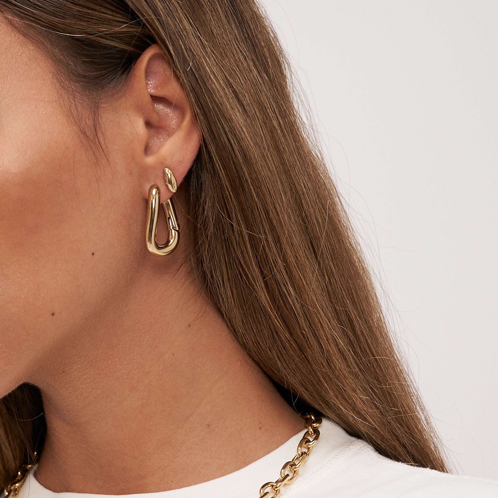 Nahaufnahme einer Frau, die Copenhagen Studios COPENHAGEN STUDIOS Ohrstecker - Organics goldene Creolen, Mini-Ohrstecker und eine goldene Halskette vor einem hellen Hintergrund trägt.