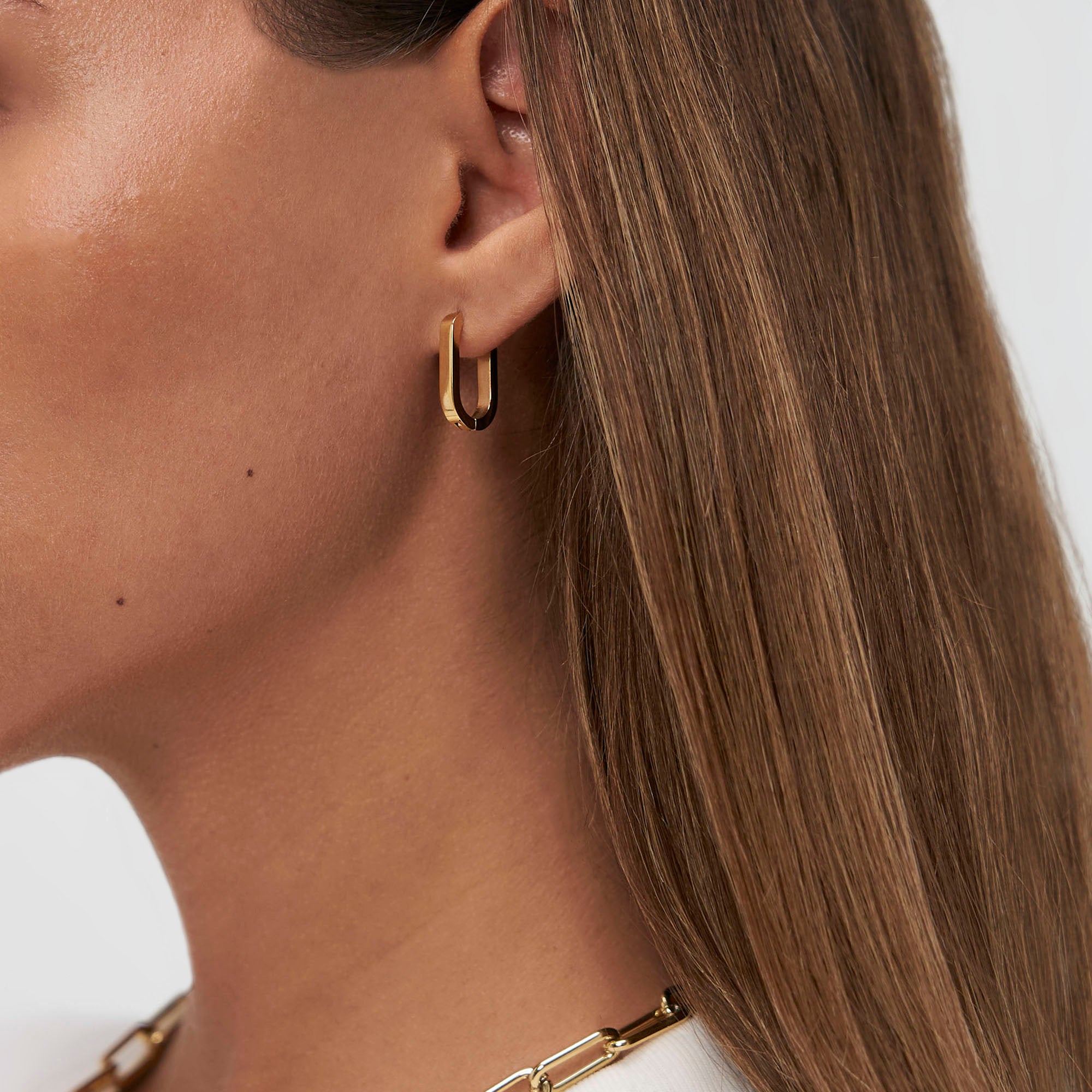 Nahaufnahme einer Frau, die die goldenen Ohrringe COPENHAGEN STUDIOS Creole - Bold Set von Copenhagen Studios und eine goldene Halskette trägt. Ihr glattes braunes Haar unterstreicht das minimalistische Design.