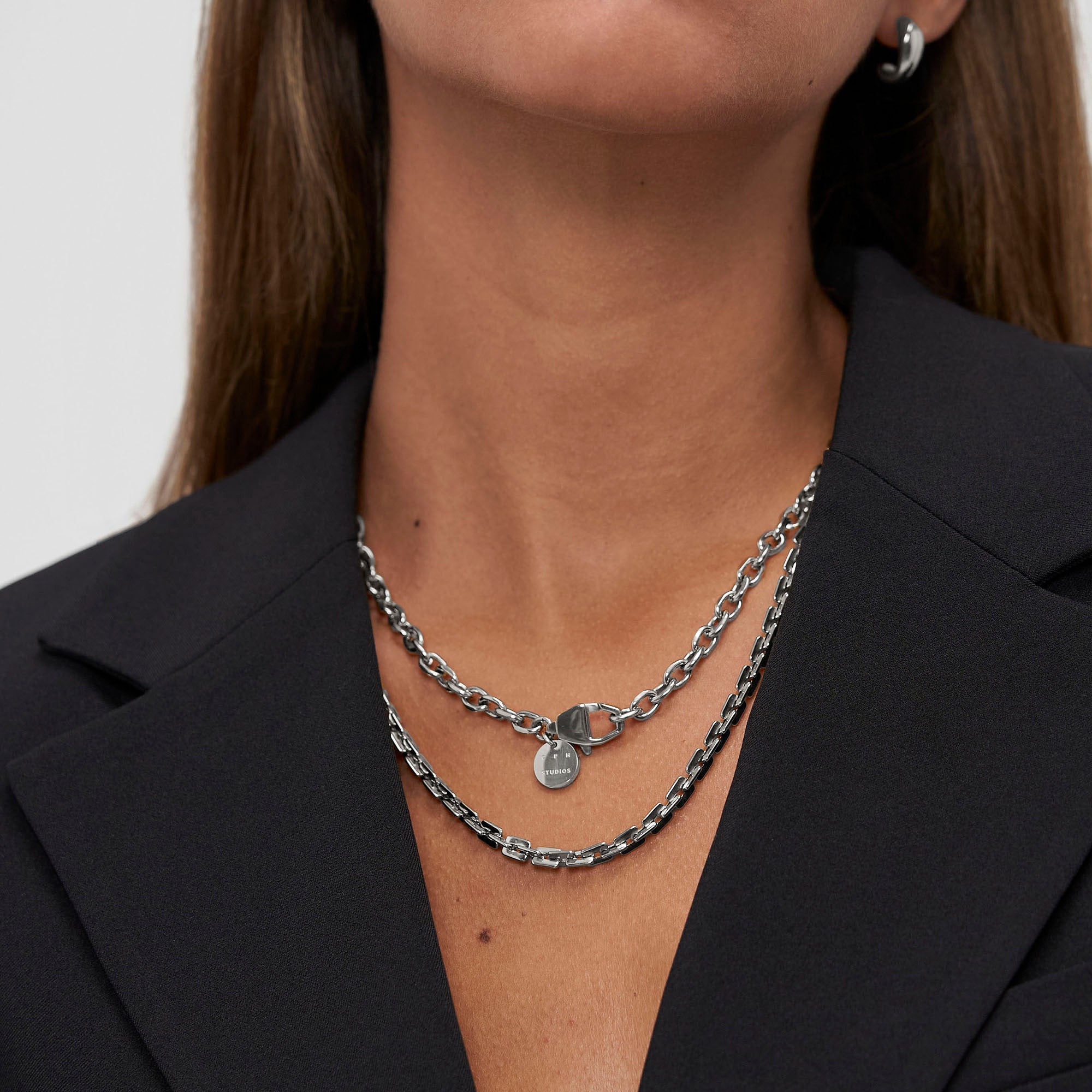 Eine Frau in einem schwarzen Blazer trägt mehrlagige Silberketten, darunter die COPENHAGEN STUDIOS Halskette - Basic Chains von Copenhagen Studios, sowie eine klobige Halskette und einen kleinen Creolenring.