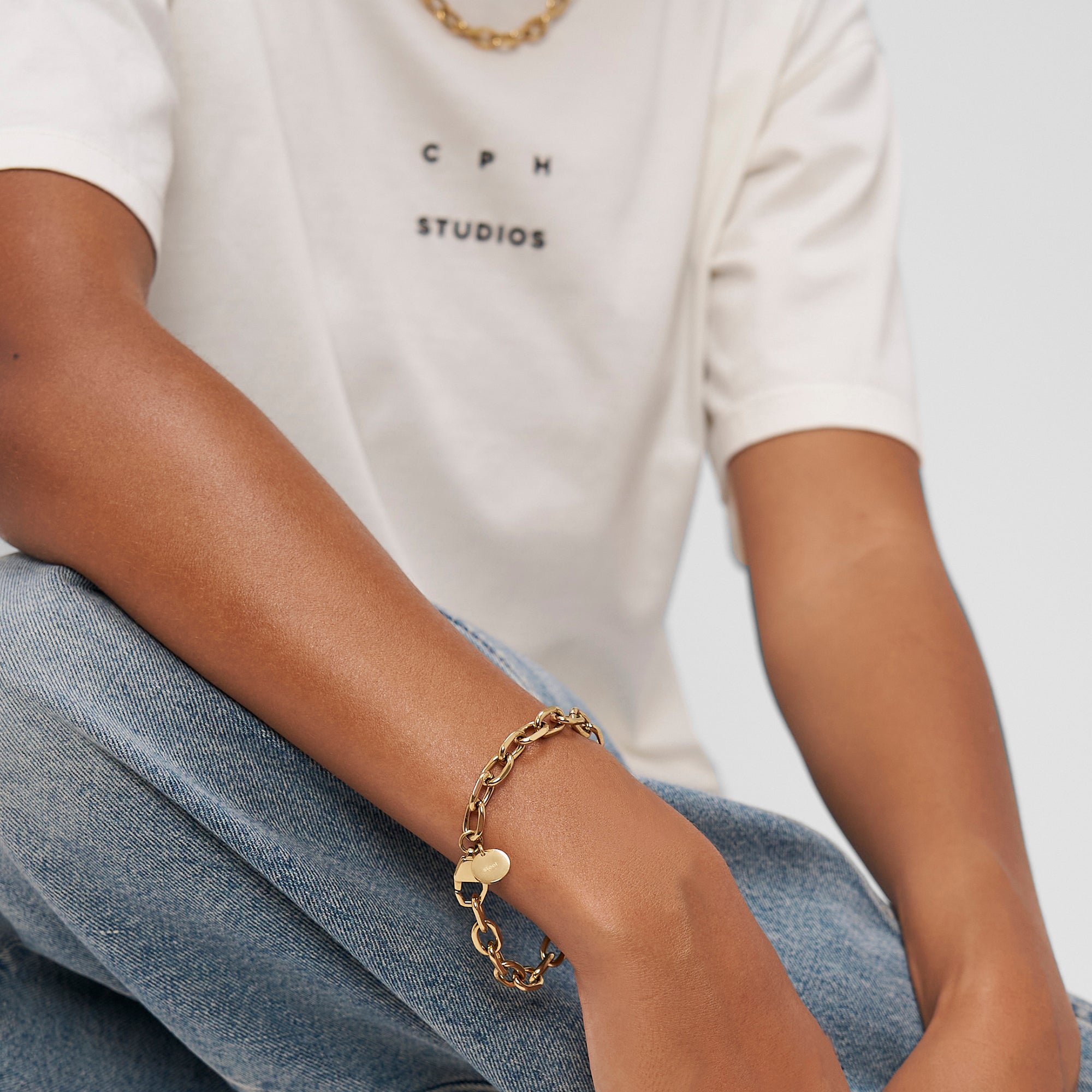 Die Person trägt ein weißes T-Shirt, blaue Jeans und goldenen Schmuck mit dem Copenhagen Studios Armband - Basic Chains - mühelos stilvoll mit dem typischen Copenhagen Studios Flair.