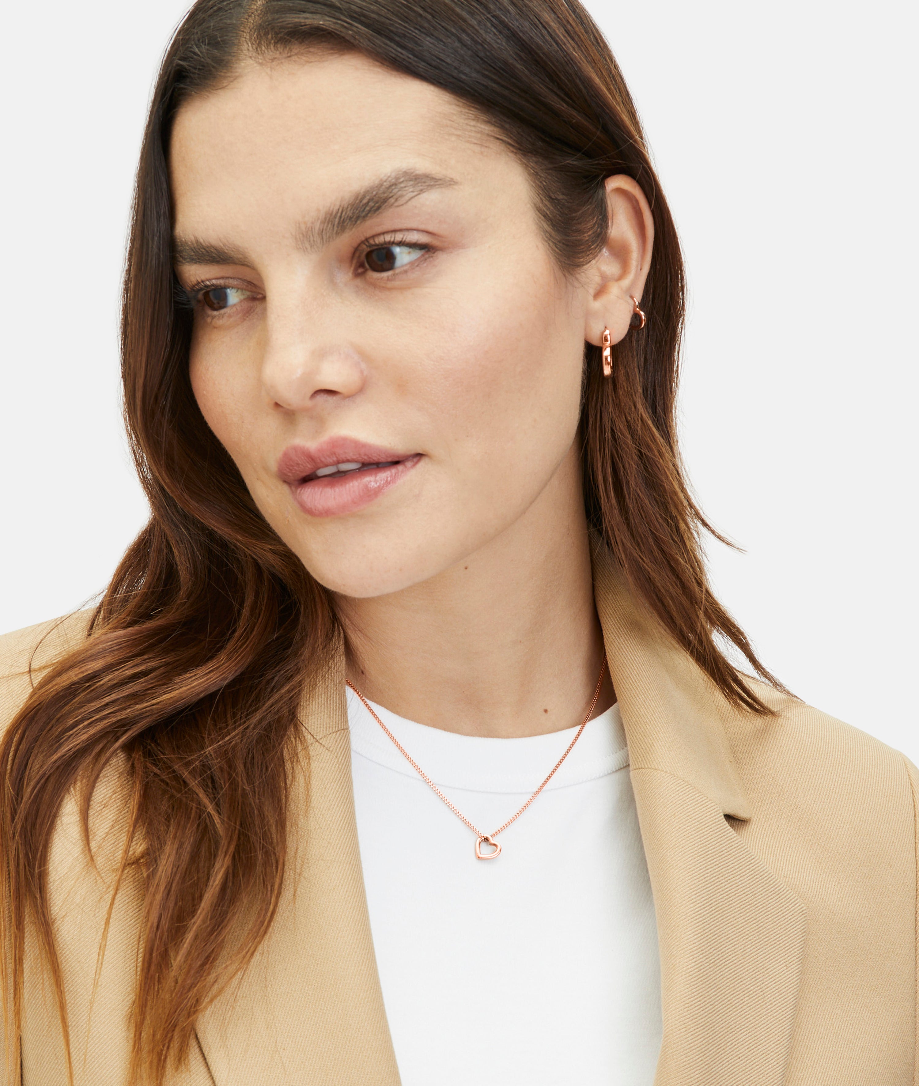 Frau mit gewelltem braunem Haar trägt einen beigen Blazer, ein weißes Oberteil, Goldschmuck und die Tamaris Halskette - The Love Heart von Tamaris mit einem herzförmigen Anhänger, mit Blick zur Seite.
