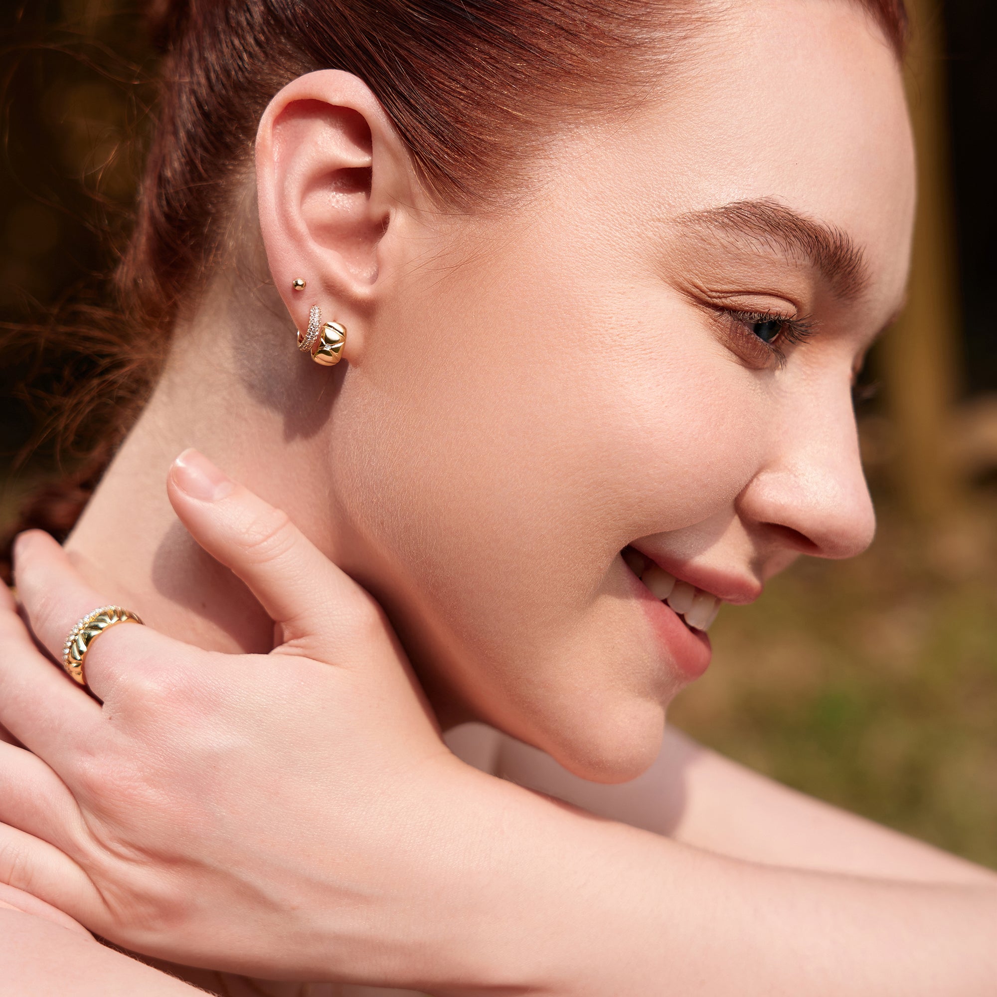 Eine Frau lächelt im Freien, sie trägt Ania Haie Ohrringe - Gold Kiss Hoops von Ania Haie und einen passenden Ring, ihre Hand berührt sanft ihren Hals.
