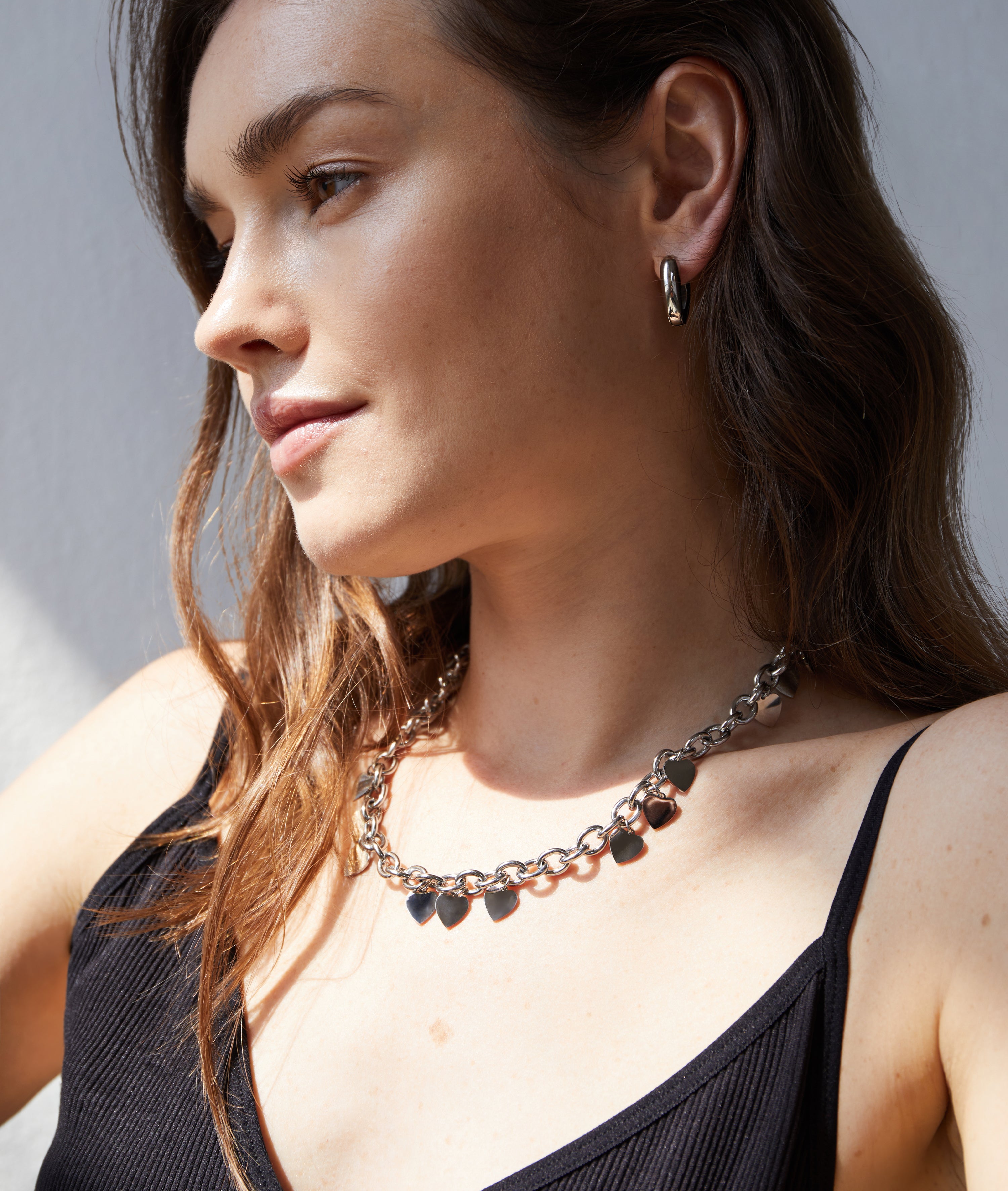 Frau mit gewelltem braunem Haar, trägt eine silberne Halskette und Echtzeit Creole - The Modern Basic von Echtzeit, gepaart mit einem schwarzen Oberteil - ein modernes Accessoire, das nach links schaut.
