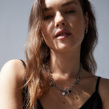Eine Frau mit gewelltem braunem Haar trägt eine silberne Herzkette, Echtzeit Creole - The Modern Basic Ohrringe von Echtzeit und ein schwarzes Oberteil im Sonnenlicht.
