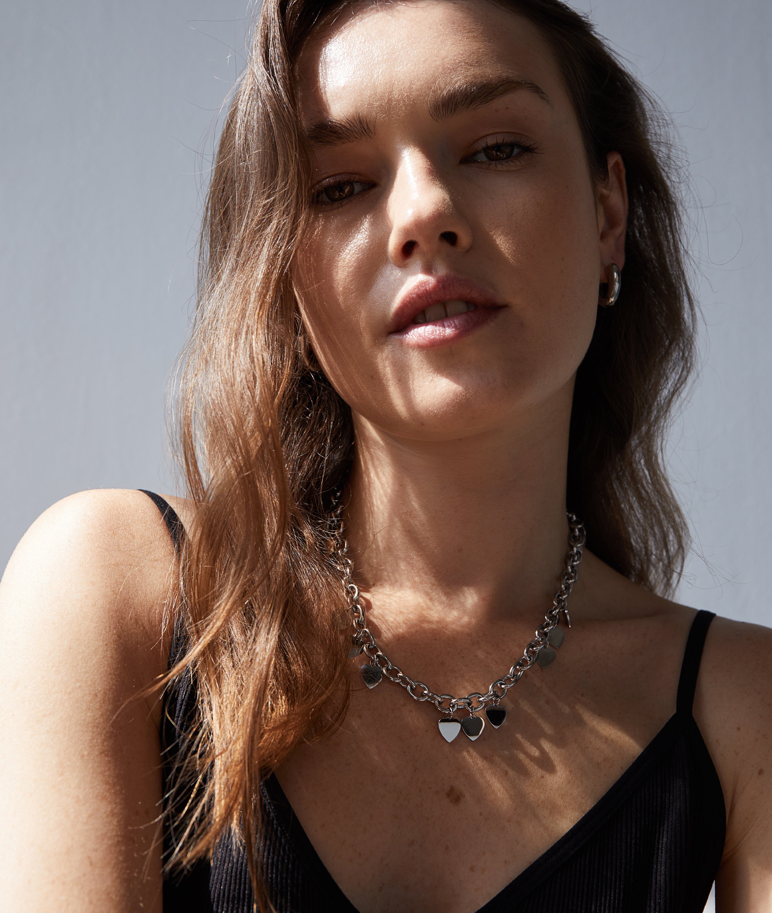 Eine Frau mit gewelltem braunem Haar trägt eine silberne Herzkette, Echtzeit Creole - The Modern Basic Ohrringe von Echtzeit und ein schwarzes Oberteil im Sonnenlicht.