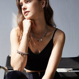 Eine Frau in einem schwarzen Oberteil sitzt auf einem Stuhl, das Sonnenlicht unterstreicht ihren nachdenklichen Blick. Sie trägt die silbernen Ohrringe "Echtzeit Creole - The Modern Basic" von Echtzeit als modernes Accessoire.