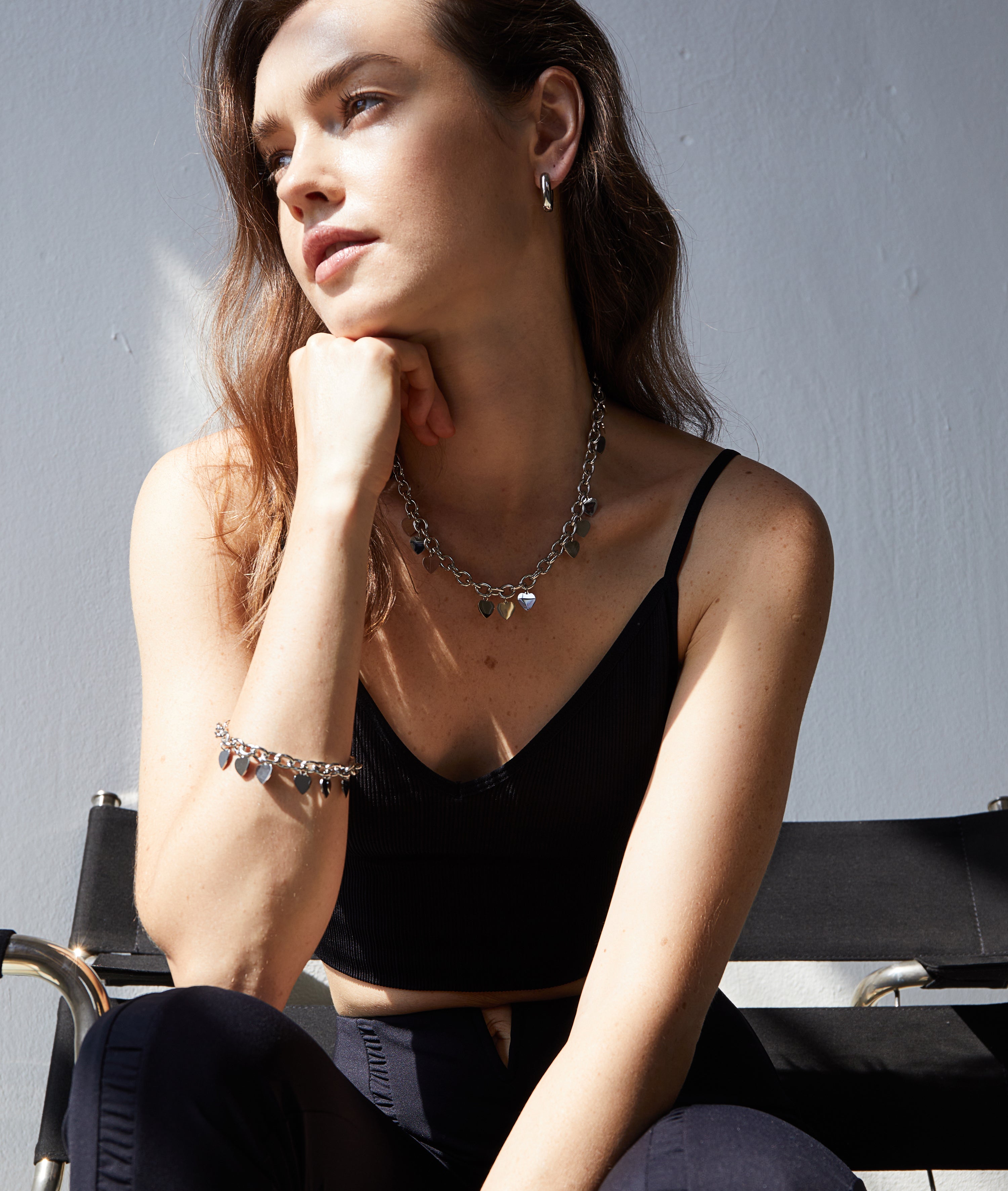 Eine Frau in einem schwarzen Oberteil sitzt auf einem Stuhl, das Sonnenlicht unterstreicht ihren nachdenklichen Blick. Sie trägt die silbernen Ohrringe "Echtzeit Creole - The Modern Basic" von Echtzeit als modernes Accessoire.