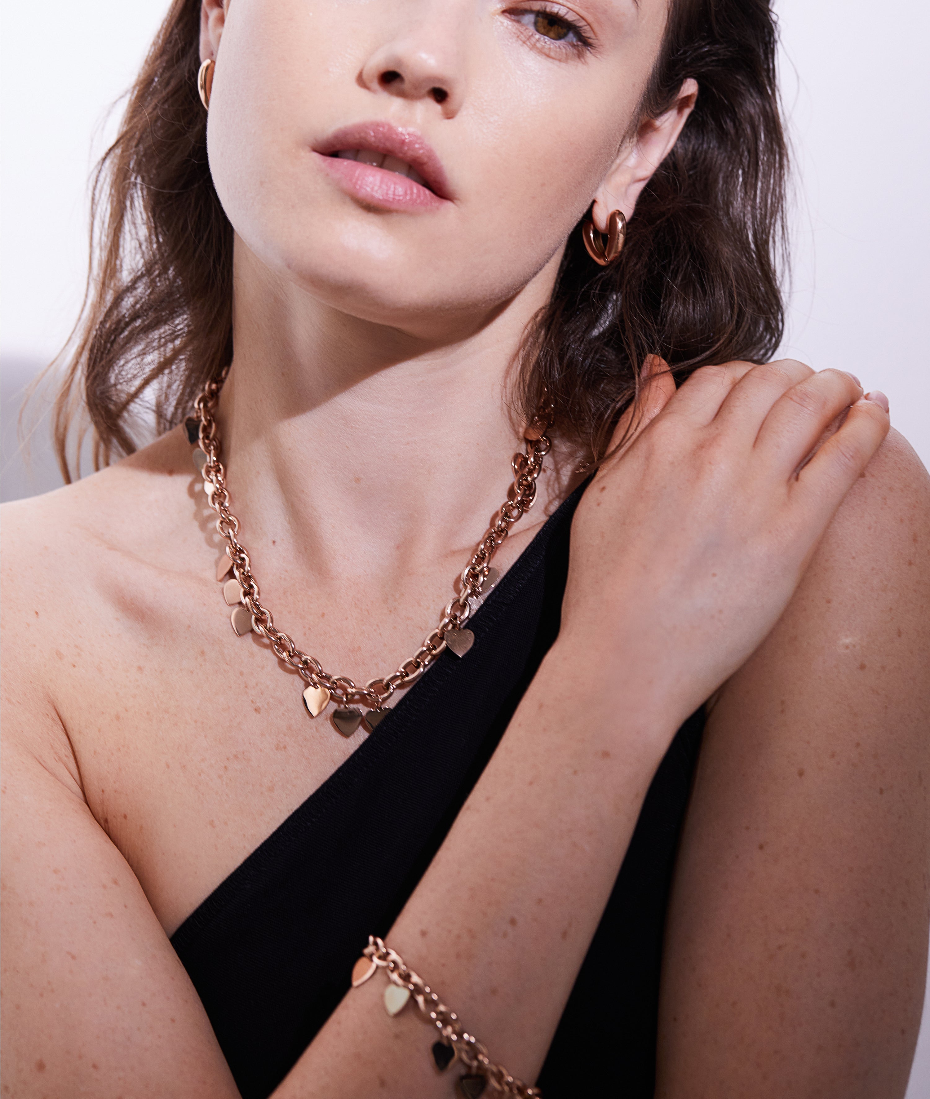 Eine Frau trägt die Echtzeit Creole - The Modern Basic von Echtzeit: ein goldenes Schmuckset mit Herz-Halskette, Armband und Statement-Ring-Ohrringen - ein modernes, elegantes Accessoire für Frauen, das perfekt zu ihrem schwarzen Outfit passt.