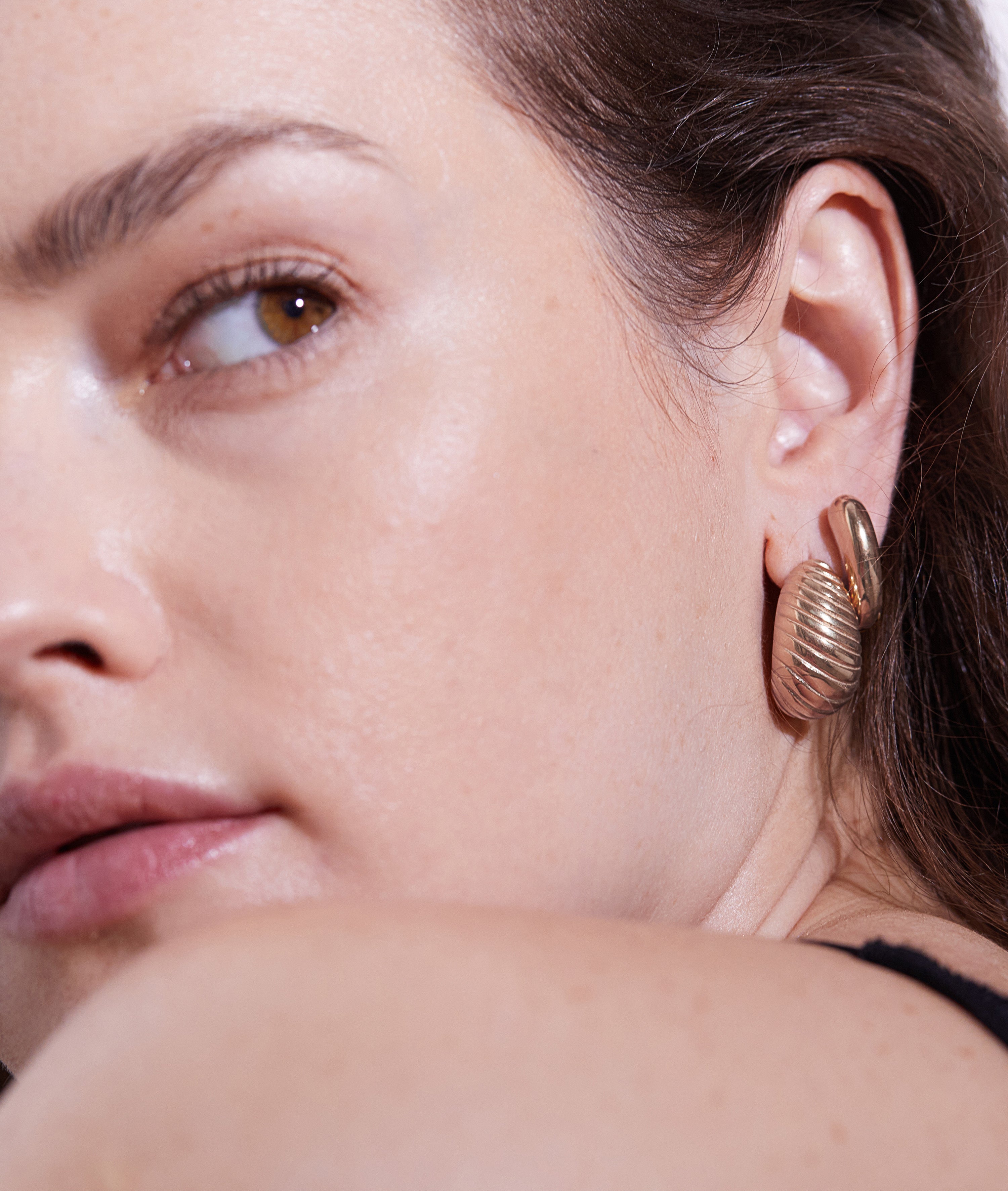 Nahaufnahme eines Frauengesichts, das ihr Ohr zeigt. Sie trägt Echtzeit Creole - The Modern Basic von Echtzeit, einen goldenen Ohrring in Muschelform - ein elegantes Accessoire für Frauen mit modernem Design.