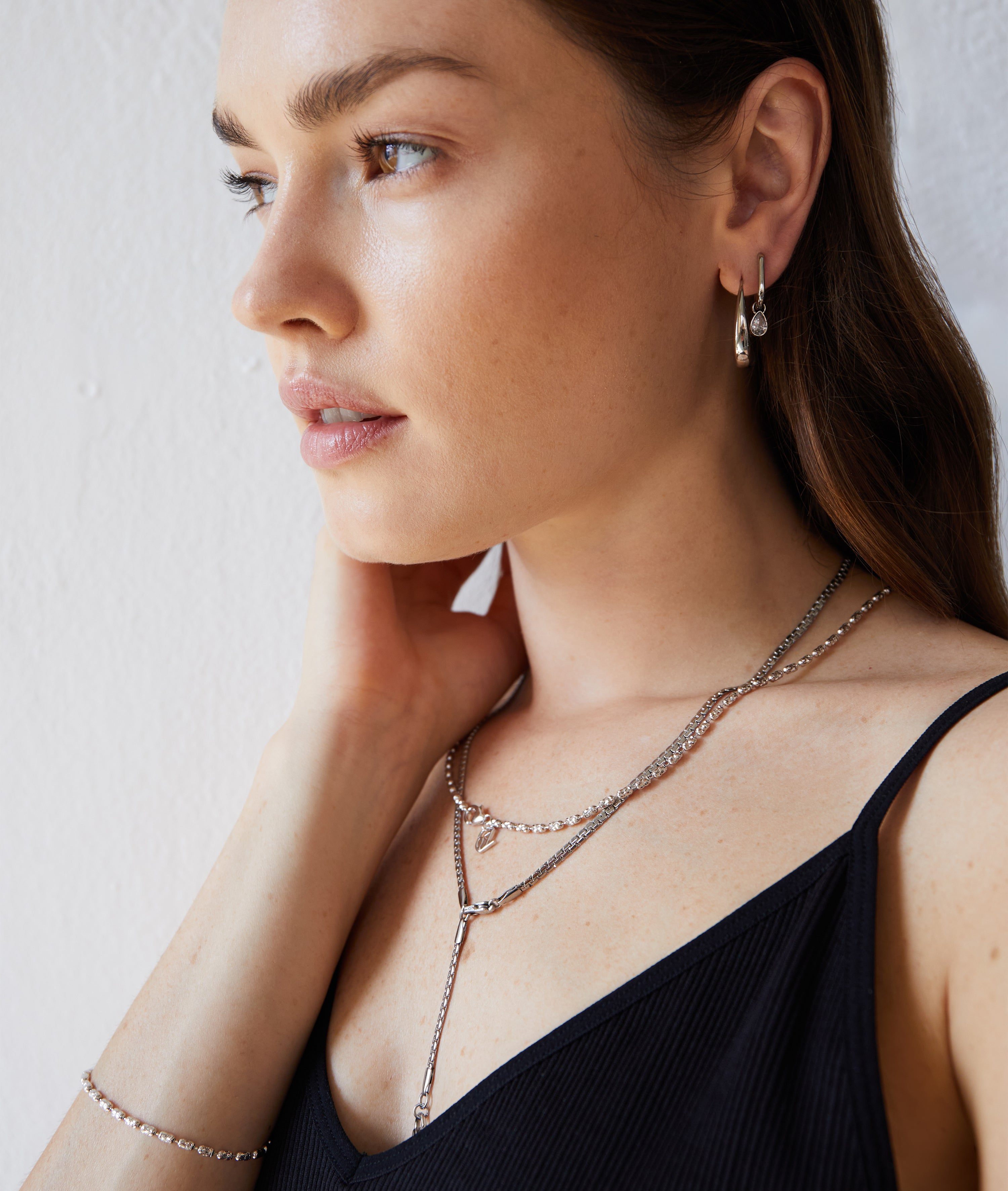 Eine Frau in einem schwarzen Oberteil trägt mehrlagigen Silberschmuck, darunter die Echtzeit Creole - Like Diamonds Ohrringe von Echtzeit, gepaart mit passenden Halsketten und einem Armband.