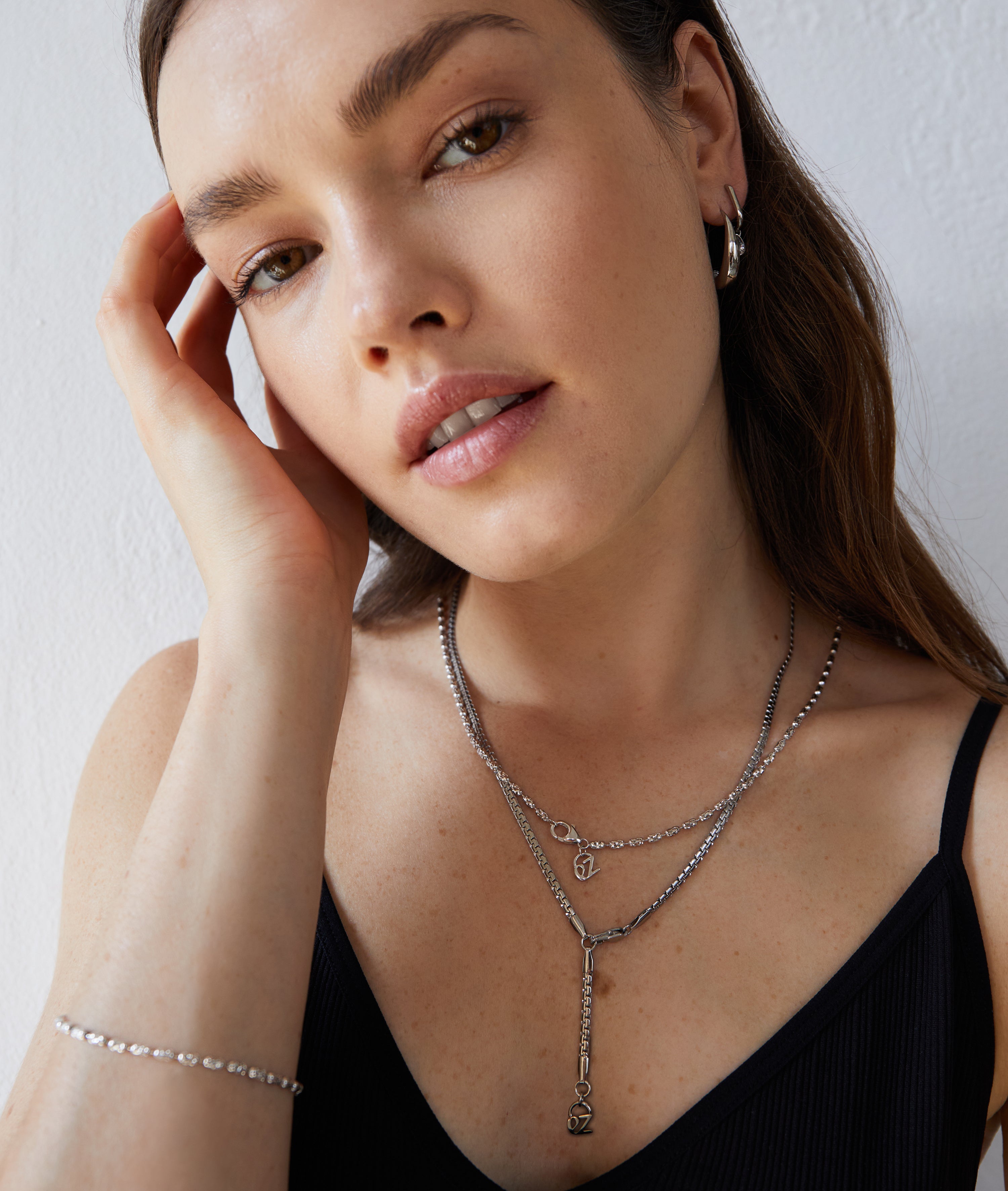 Frau mit langen braunen Haaren trägt Echtzeit Creole - Like Diamonds Silberohrringe von Echtzeit und ein schlichtes schwarzes Oberteil. Sie posiert mit der Hand neben ihrem Gesicht.