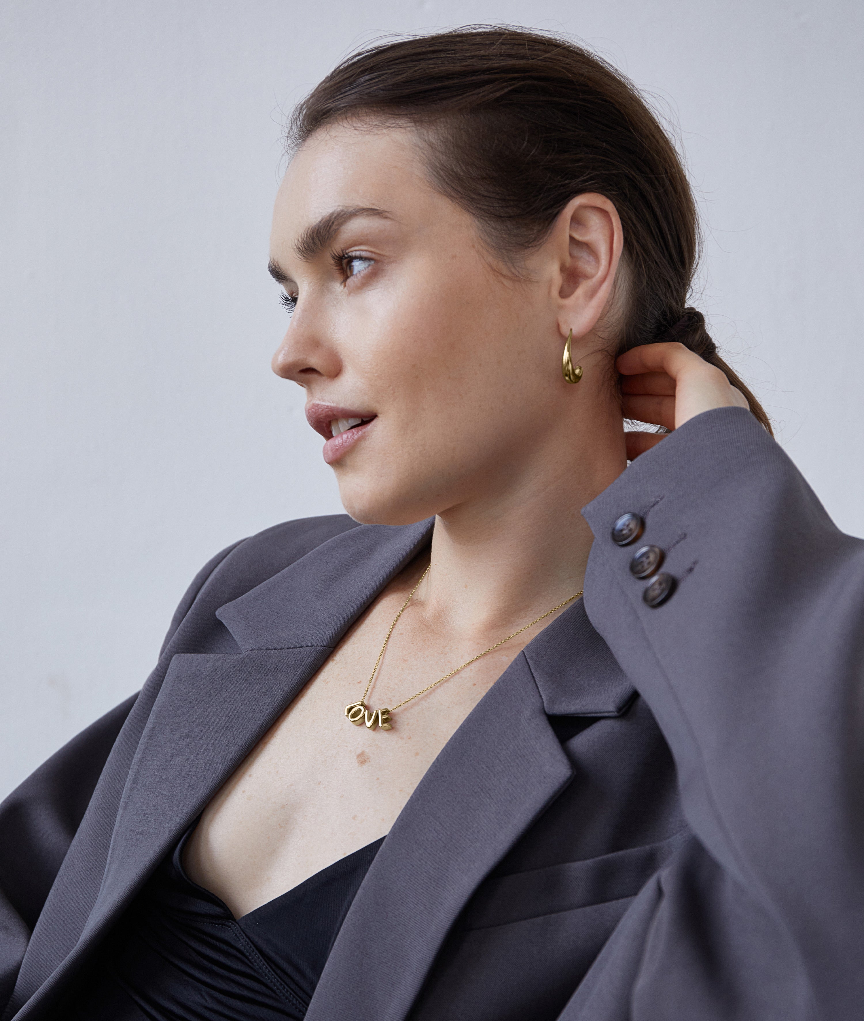 Eine Frau in einem grauen Blazer und mit Goldschmuck trägt die Echtzeit Halskette - Love Letters by Echtzeit und schaut zur Seite, während sie mit der Hand ihr Haar berührt.