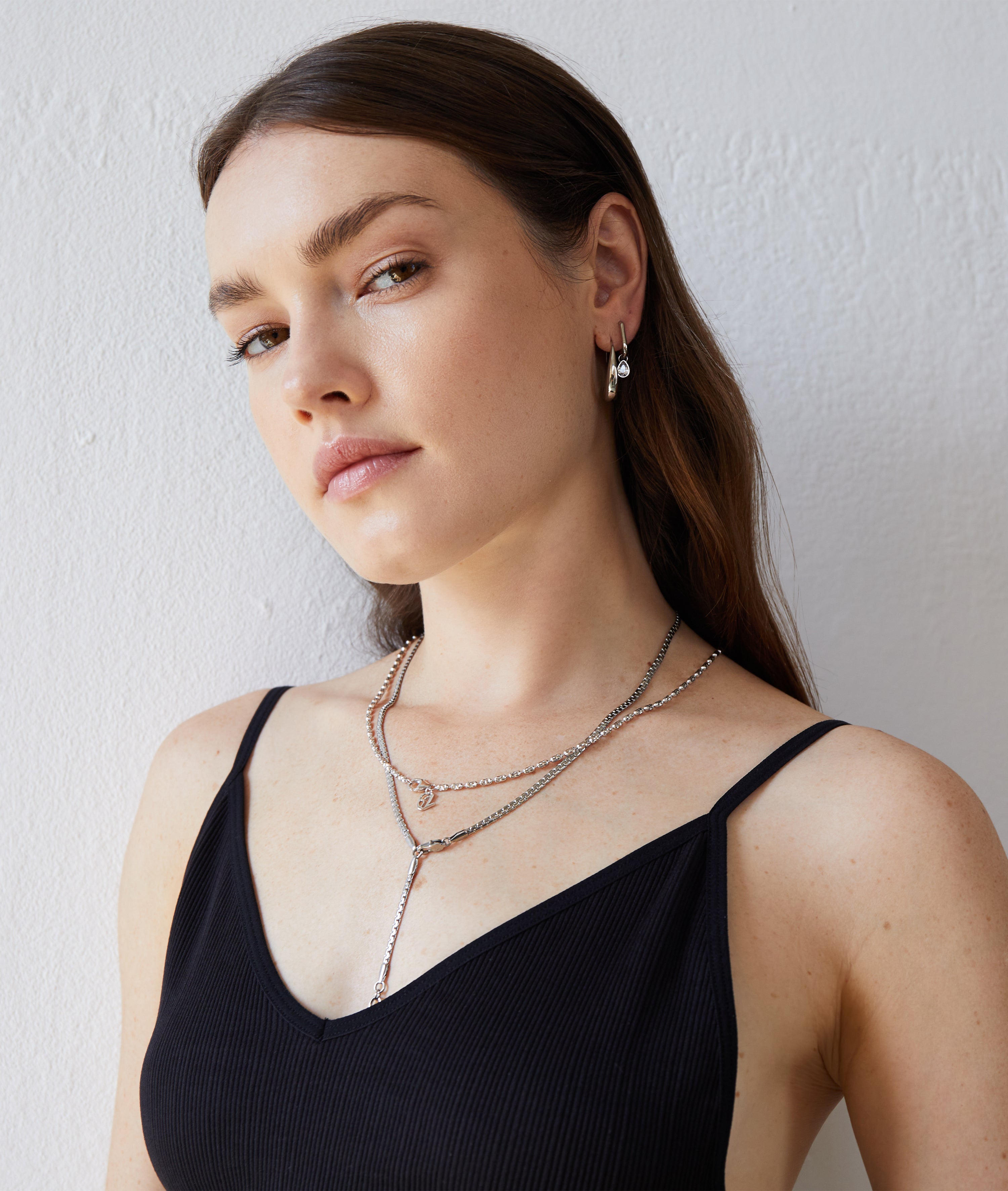 Frau mit langen braunen Haaren trägt die Echtzeit Halskette - The Sleek One von Echtzeit, kombiniert mit Ohrringen und einem schwarzen Tanktop, vor einer weißen Wand stehend.
