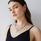 Frau mit langen braunen Haaren, die mehrlagige Silberketten, die Echtzeit Creole - Like Diamonds Ohrringe von Echtzeit, Ohrringe und ein schwarzes ärmelloses Oberteil trägt.