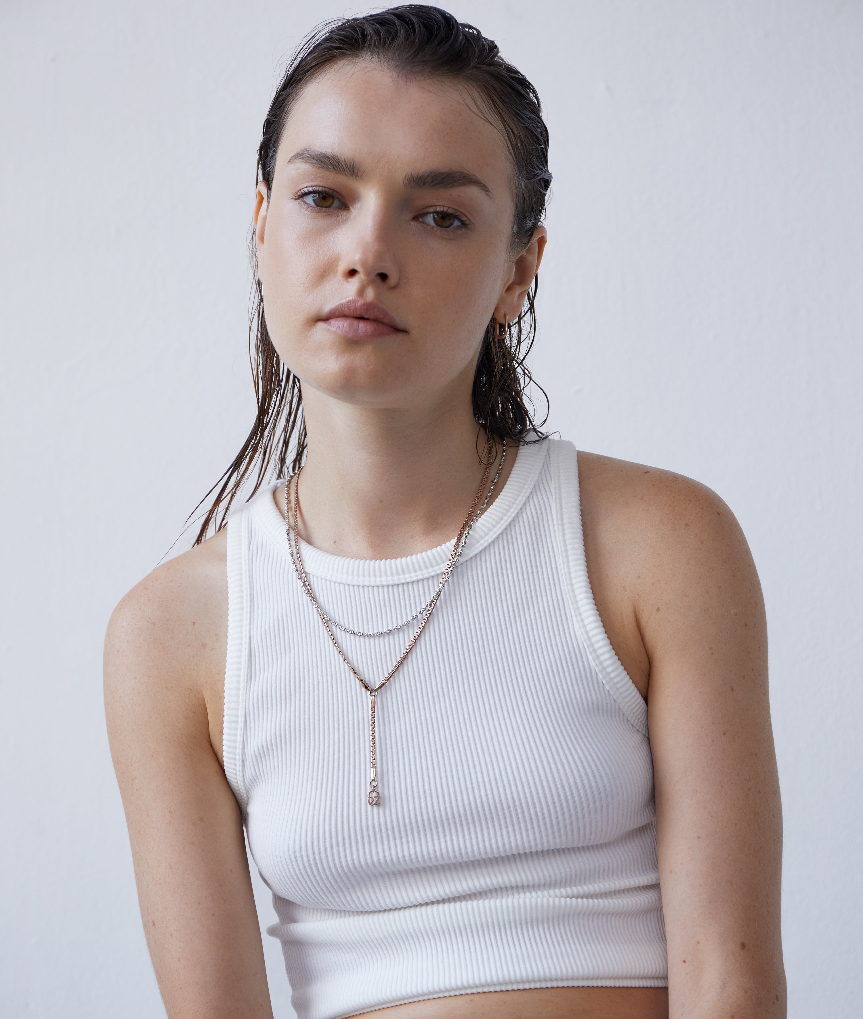 Eine Frau mit nassen Haaren trägt ein weißes geripptes Tanktop mit der Echtzeit Halskette - The Sleek One von Echtzeit, gestylt vor einem schlichten Hintergrund.