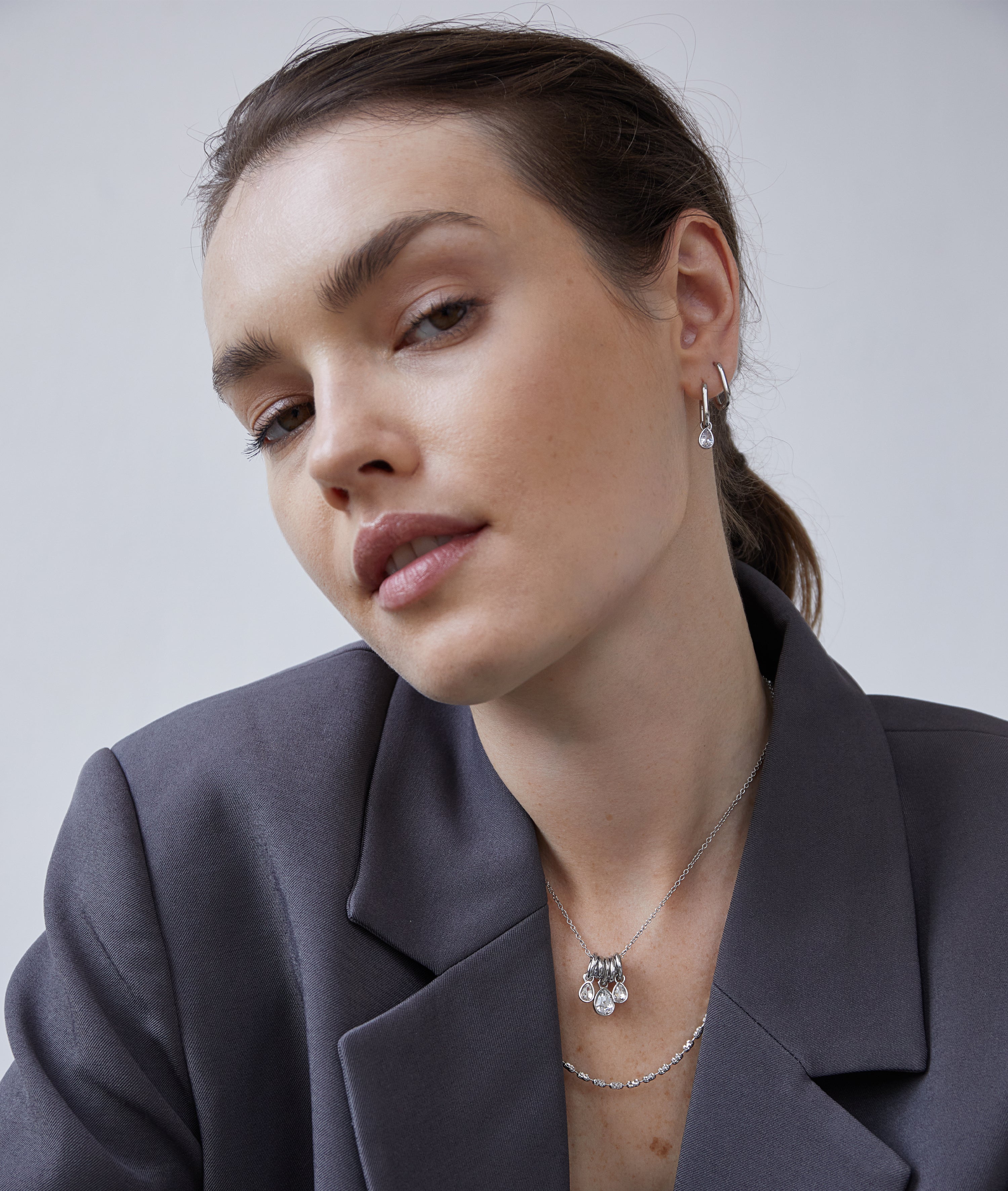 Eine Frau in einem grauen Blazer trägt die Echtzeit Creole - Like Diamonds Silberohrringe und eine mehrlagige Halskette von Echtzeit und blickt selbstbewusst in die Kamera.