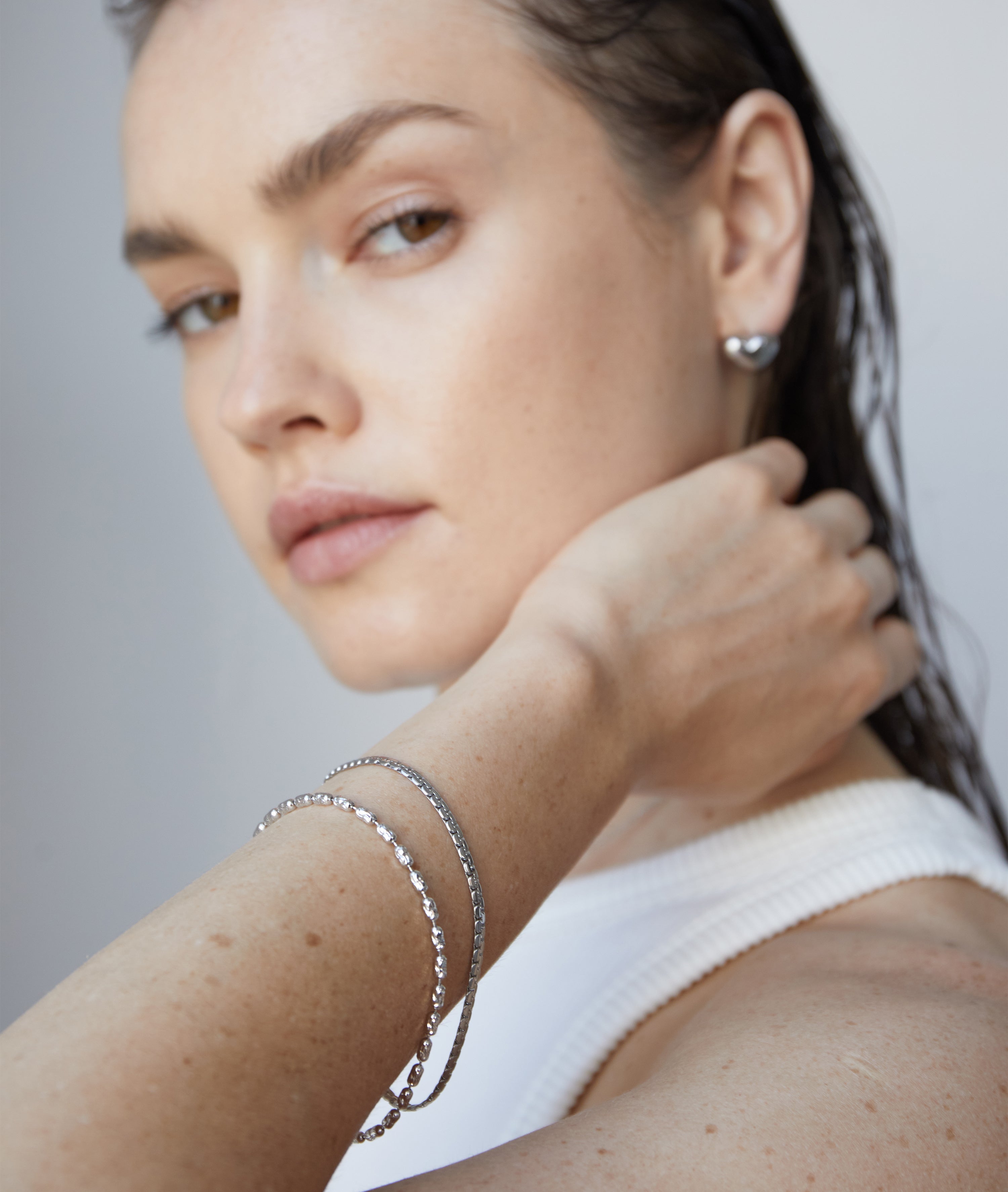 Frau mit nassen Haaren und natürlichem Make-up trägt das Echtzeit Armband - The Sleek One und silberne Ohrringe von Echtzeit, gestylt mit einem weißen Oberteil.