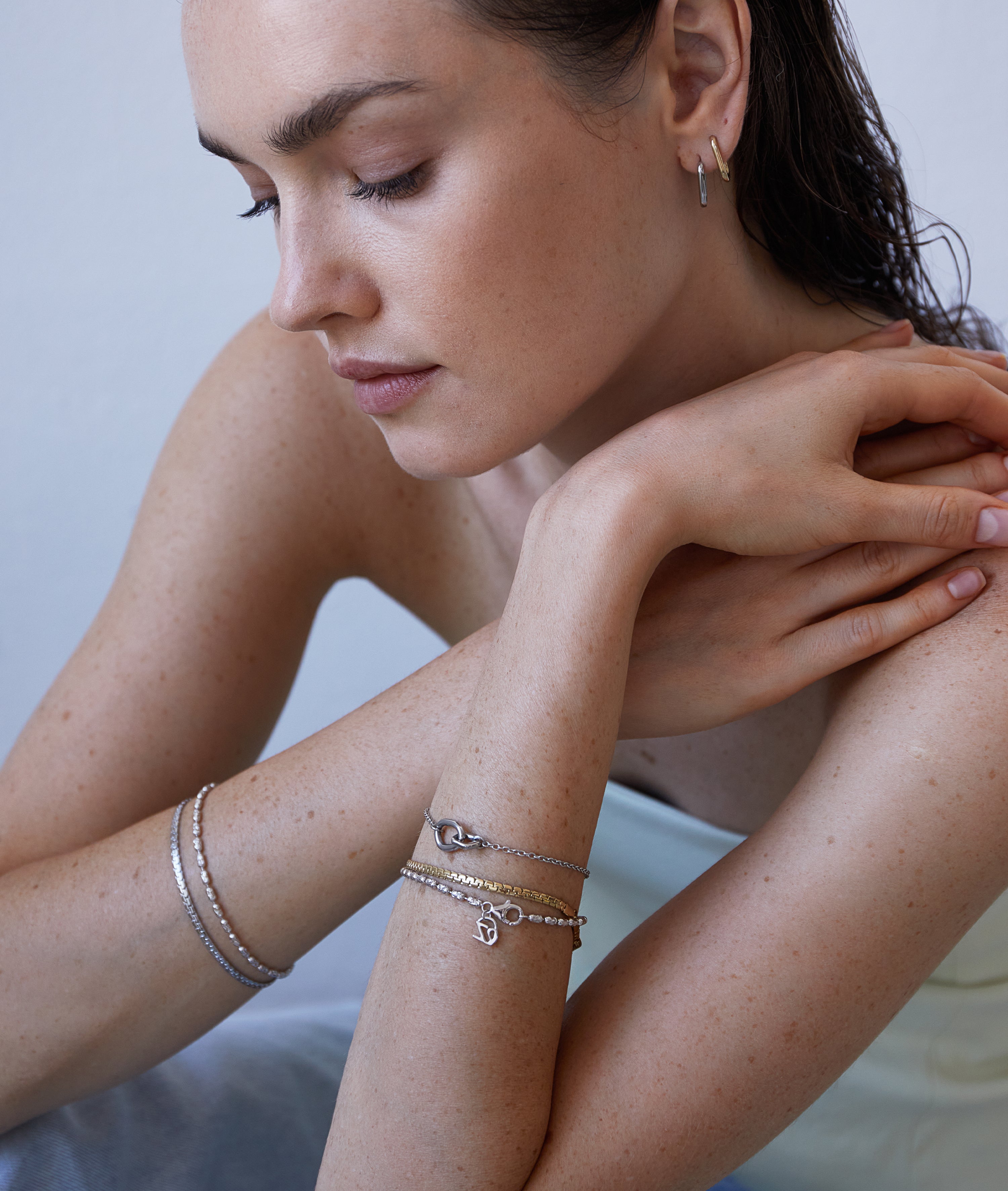 Hellhäutige Frau trägt Echtzeit Creole - Like Diamonds Silberarmbänder und Ohrringe, verschränkt die Arme und schaut nach unten.