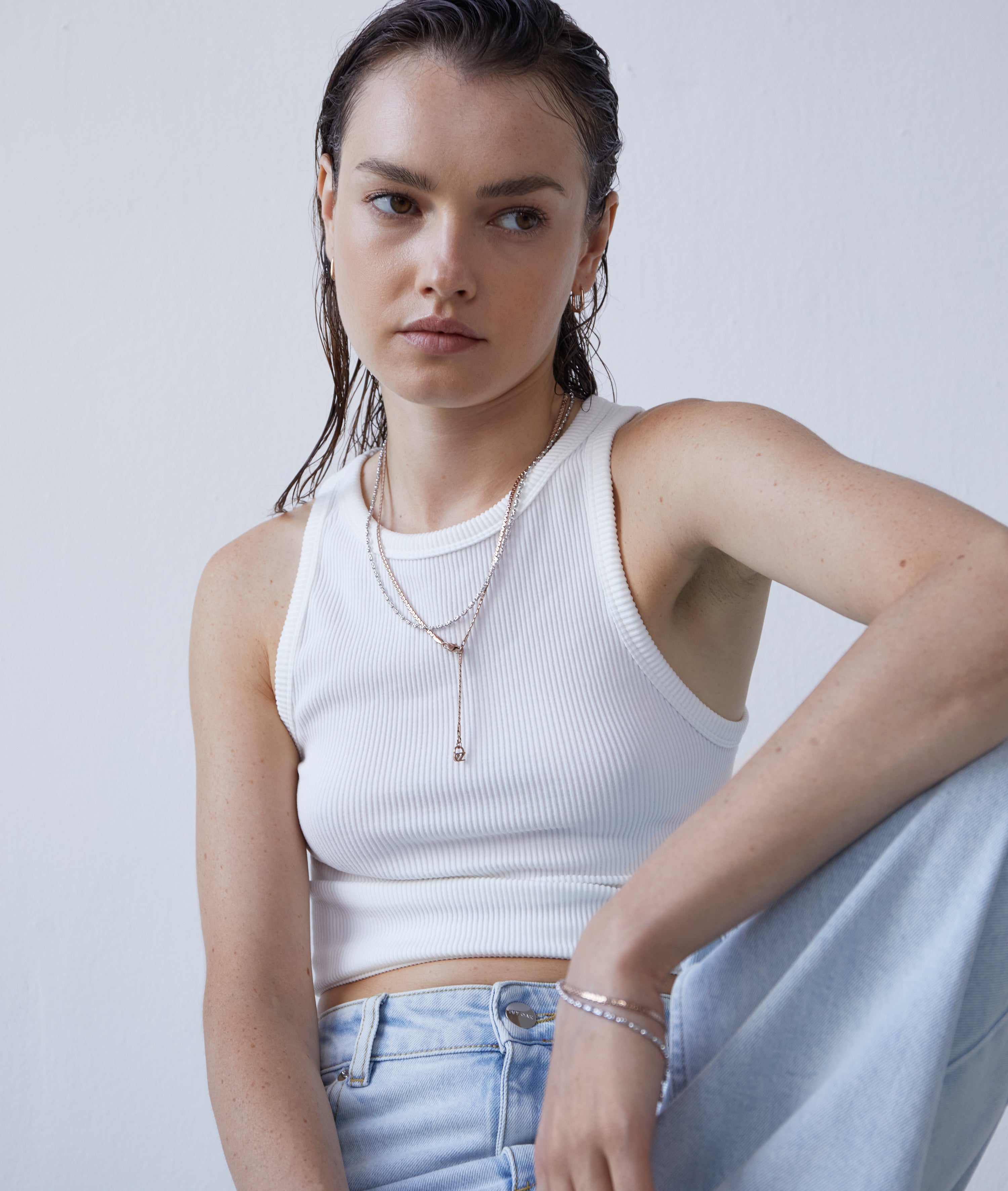 Frau mit nassen Haaren, weißem Tank-Top, hellblauen Jeans und mehrlagigen Ketten trägt das Echtzeit Armband - The Sleek One von Echtzeit und kreiert damit einen schicken Layering-Look, während sie vor einer schlichten Wand sitzt.