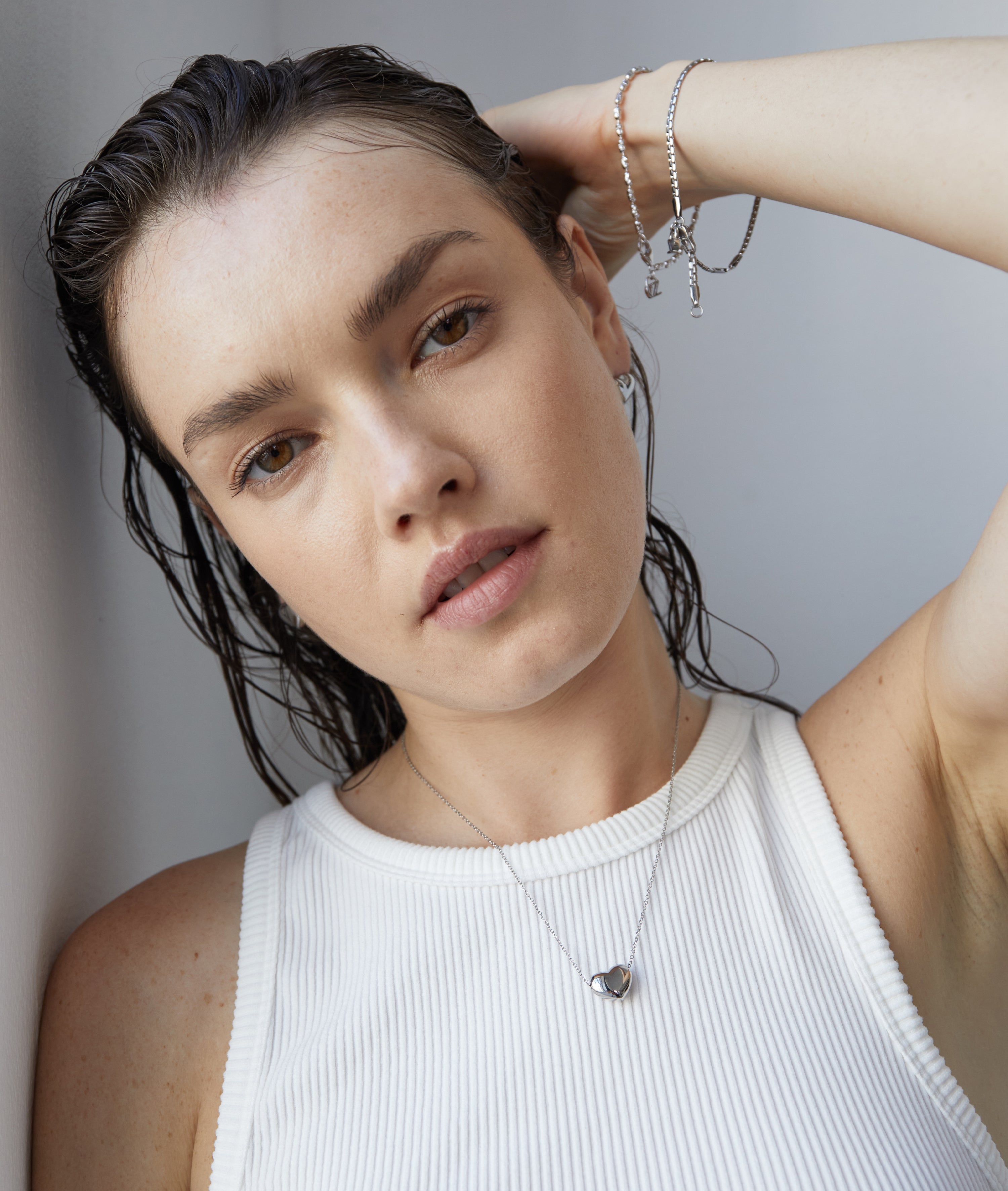 Eine Frau mit nassen Haaren in einem weißen ärmellosen Oberteil präsentiert eine Herz-Halskette und das Echtzeit Armband - The Sleek One von Echtzeit und kreiert einen schicken Lagen-Look, während sie in einem Haus posiert.