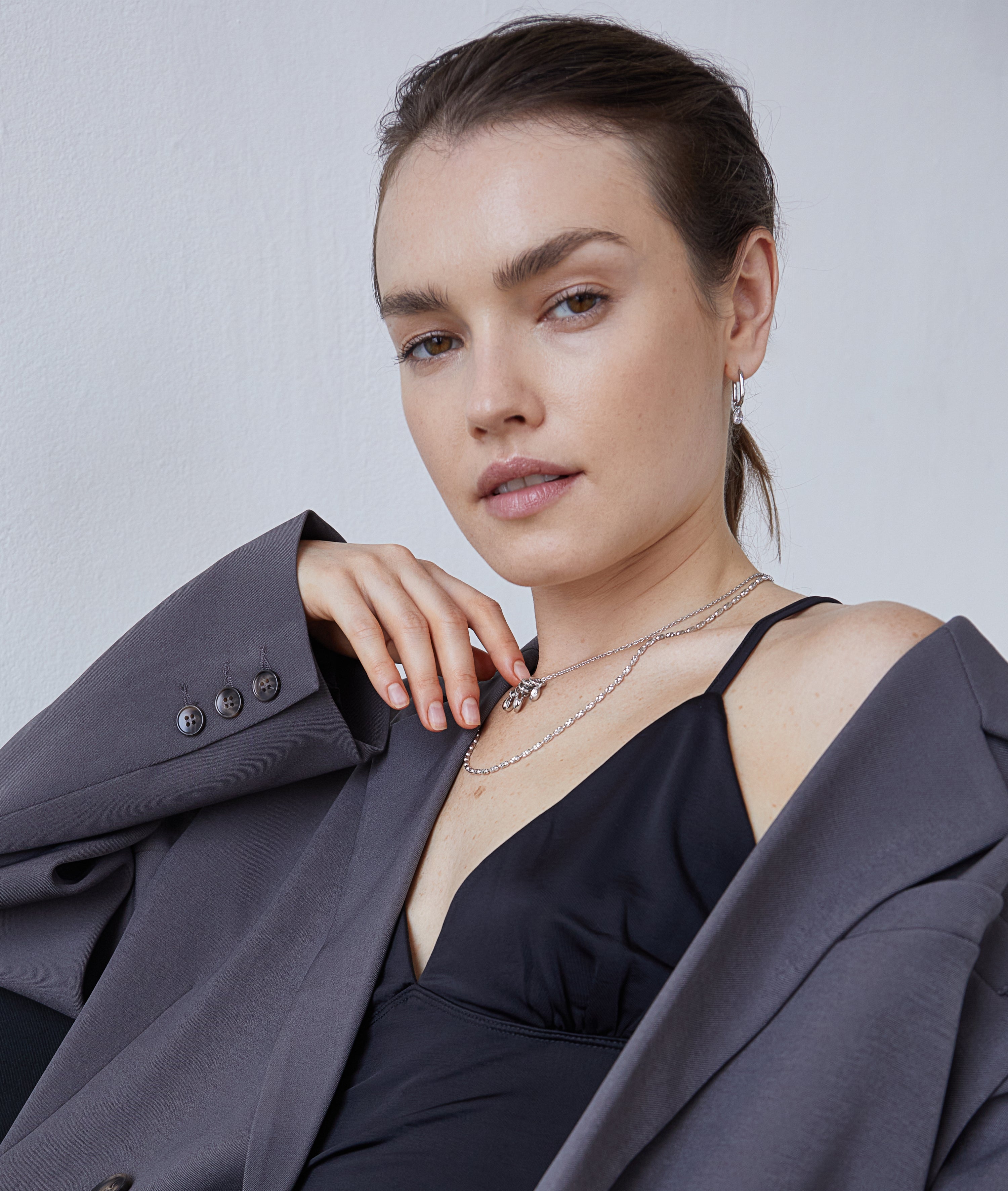 Eine Frau in einem schwarzen Oberteil und einem grauen Blazer, die mehrlagige Ketten und den Echtzeit Creole - Like Diamonds Ohrring von Echtzeit trägt, blickt selbstbewusst in die Kamera.