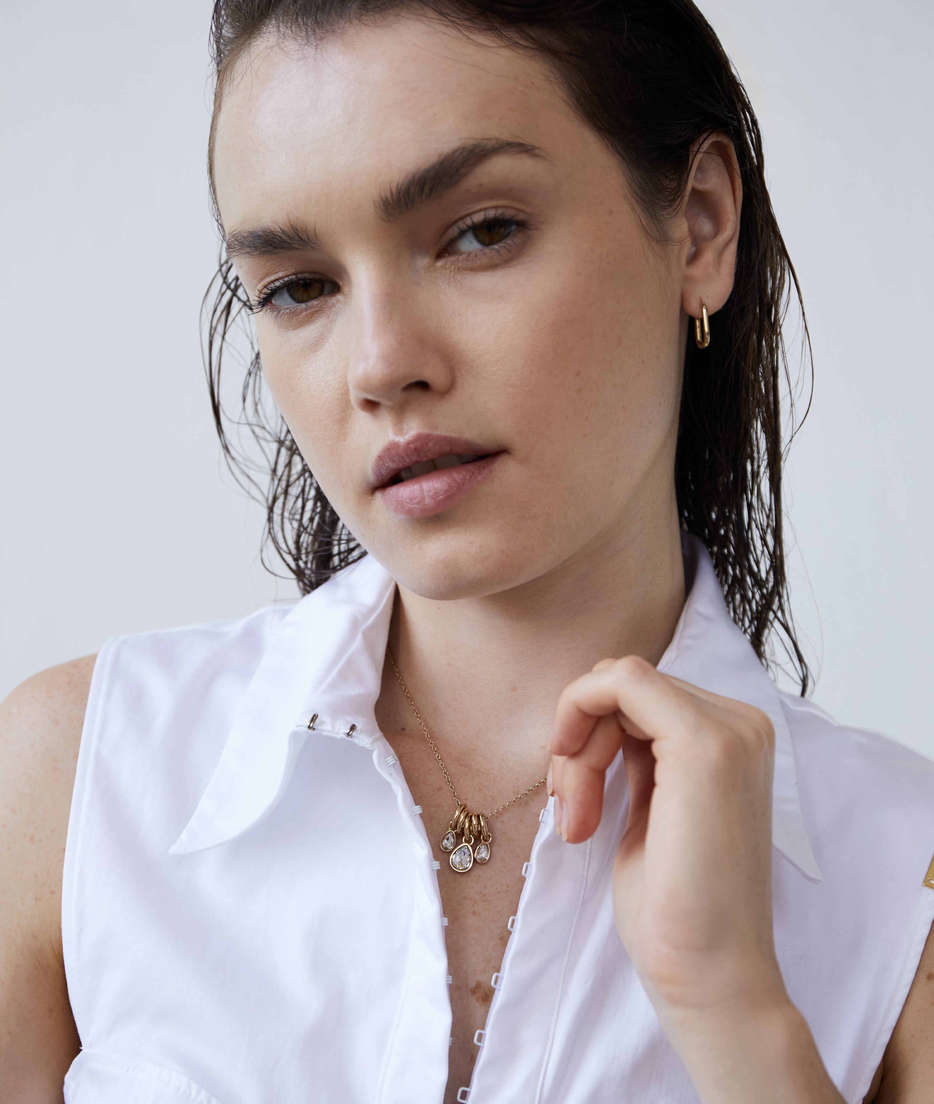 Eine Frau mit nassen Haaren in einem weißen ärmellosen Shirt trägt die Echtzeit Creole - Like Diamonds Goldohrringe von Echtzeit und geschichtete Goldketten, darunter einen zarten Tropfenanhänger.