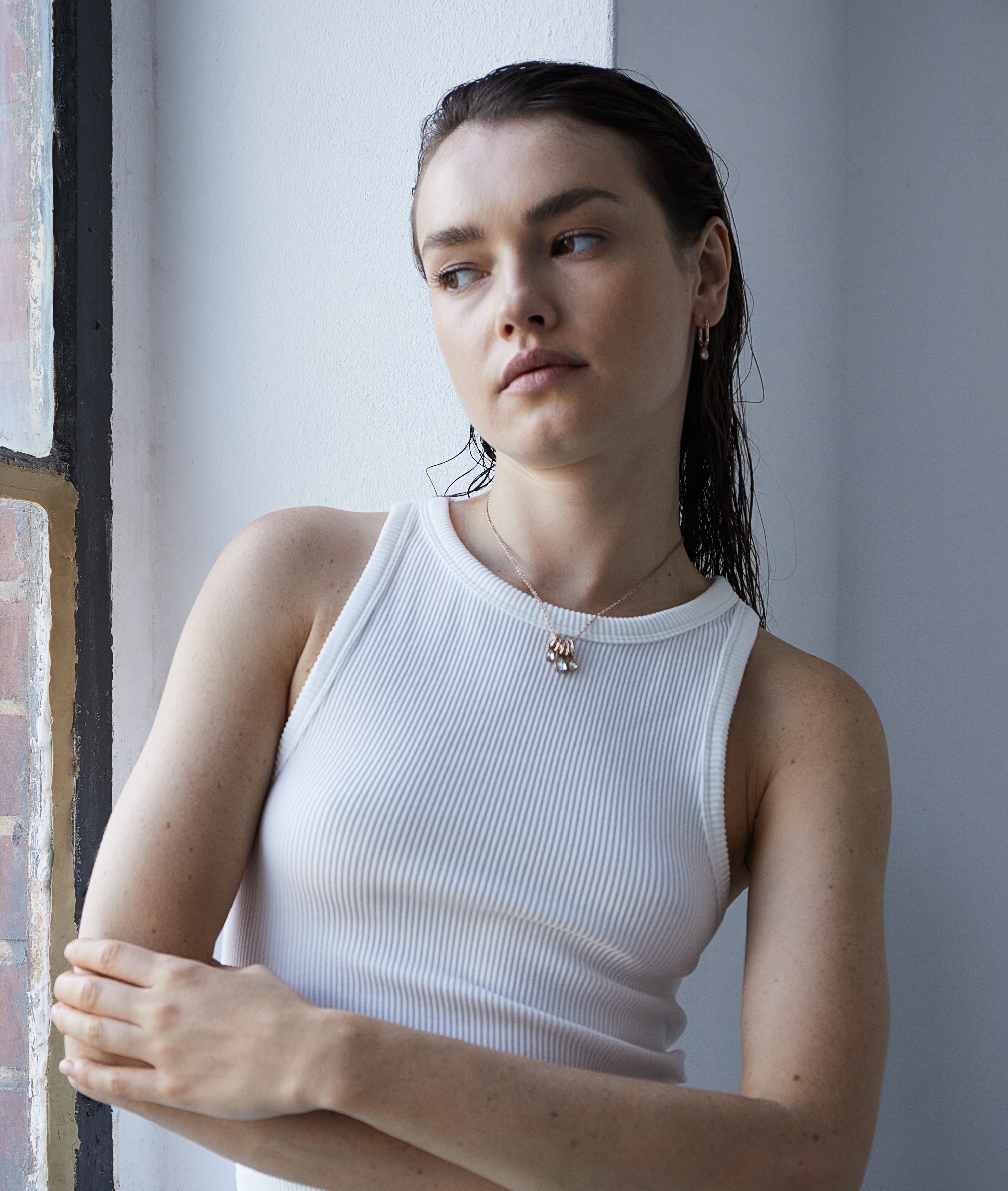 Eine Frau in einem weißen Tank-Top steht an einem Fenster und schaut mit verschränkten Armen nachdenklich zur Seite. Ihre Echtzeit Creole - Like Diamonds Ohrringe fangen das sanfte Licht ein.