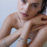 Eine Frau mit braunen Haaren stützt ihren Kopf auf ihre Arme und präsentiert das Echtzeit Armband - Like Diamonds von Echtzeit; gepaart mit Ohrringen wird dieser elegante Damen Schmuck Look durch silberne Details und ein Unendlichkeitszeichen-Motiv akzentuiert.
