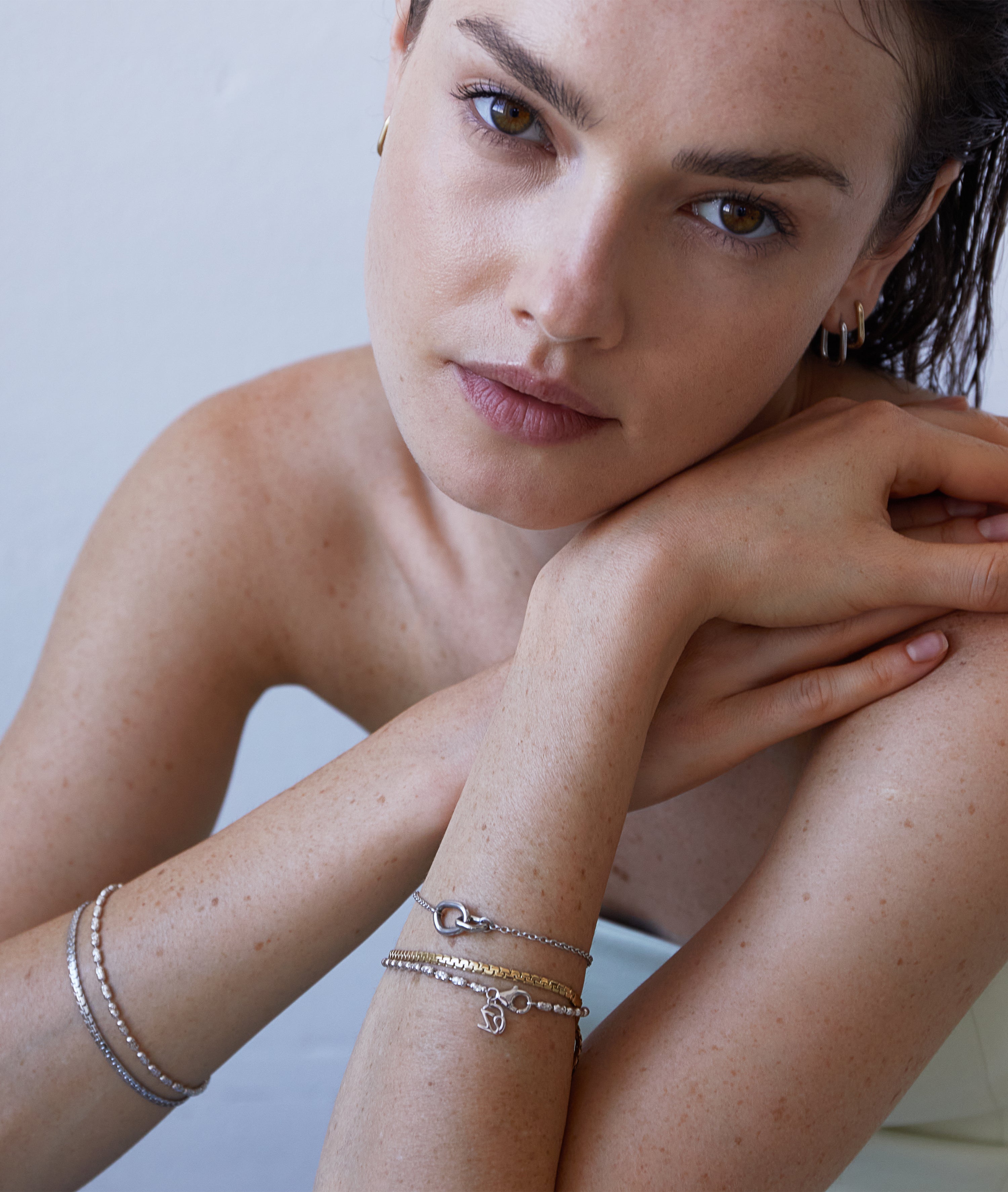 Eine Frau mit braunen Haaren stützt ihren Kopf auf ihre Arme und präsentiert das Echtzeit Armband - Like Diamonds von Echtzeit; gepaart mit Ohrringen wird dieser elegante Damen Schmuck Look durch silberne Details und ein Unendlichkeitszeichen-Motiv akzentuiert.