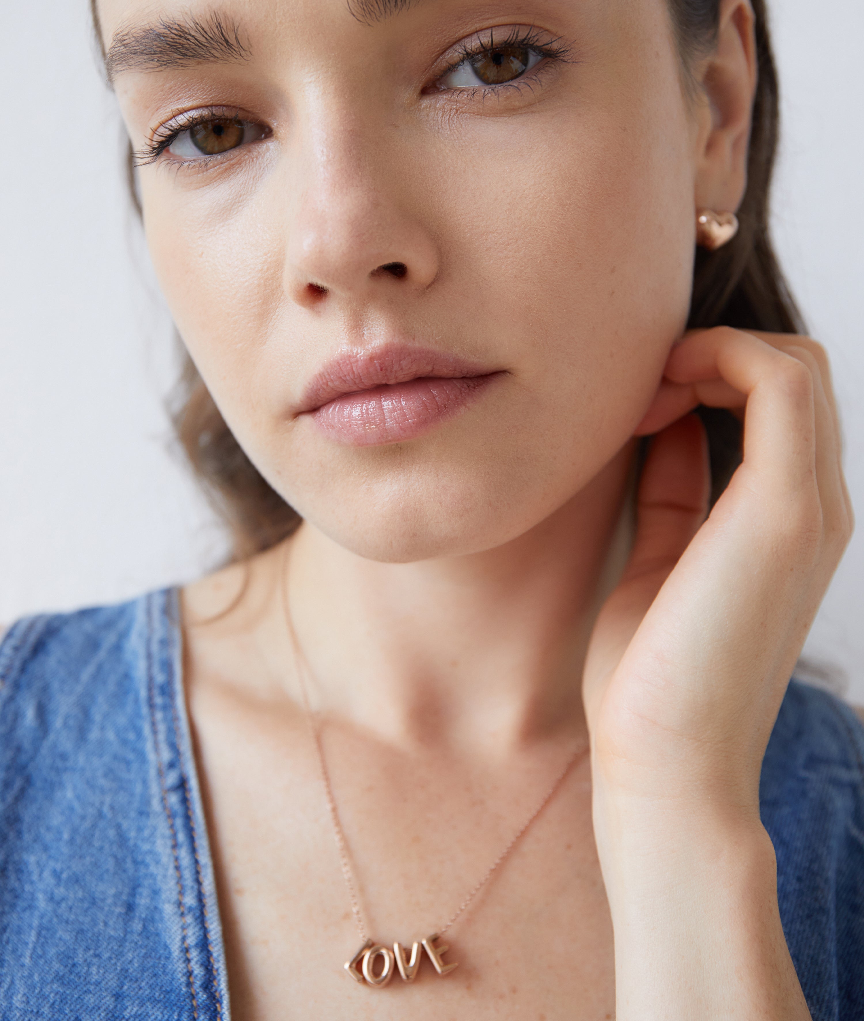 Frau trägt Echtzeit Ohrstecker - Puffy Heart von Echtzeit und ein Denim-Top, berührt ihr Gesicht und blickt sanft in die Kamera - ein Look mit zeitloser Eleganz und romantischer Note.
