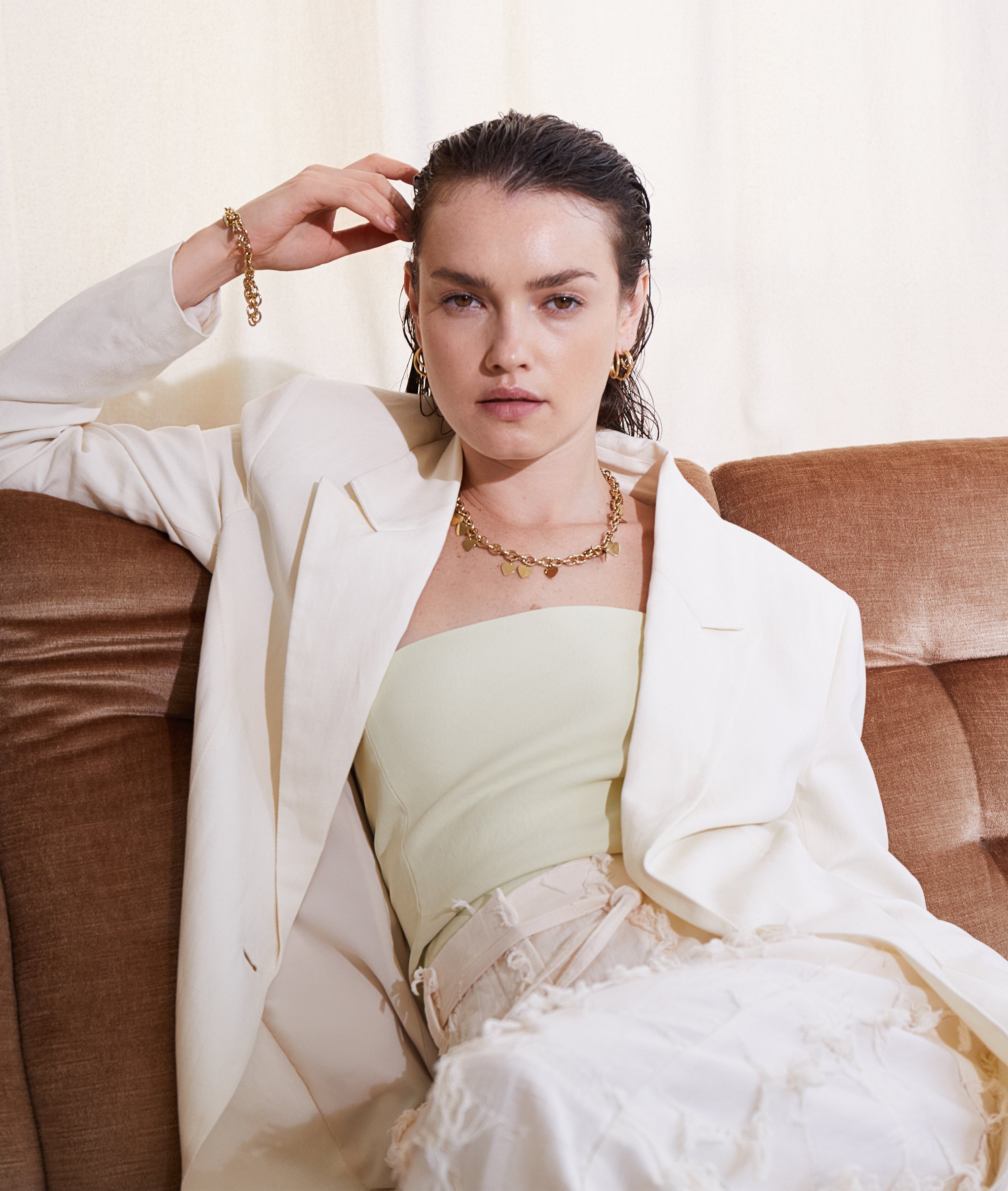 Eine Frau in weißem Blazer und goldenem Schmuck, darunter die Echtzeit Halskette - Endless Love by Echtzeit mit einem zarten Herzanhänger, sitzt auf einer braunen Couch und blickt selbstbewusst in die Kamera.