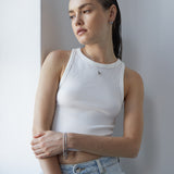 Eine Frau in einem weißen Tanktop und hellen Jeans steht an einer weißen Wand und blickt nachdenklich zur Seite. Sie trägt die Echtzeit Halskette - Puffy Heart von Echtzeit, die ihrem Look einen dezenten, zeitlosen Glanz verleiht.