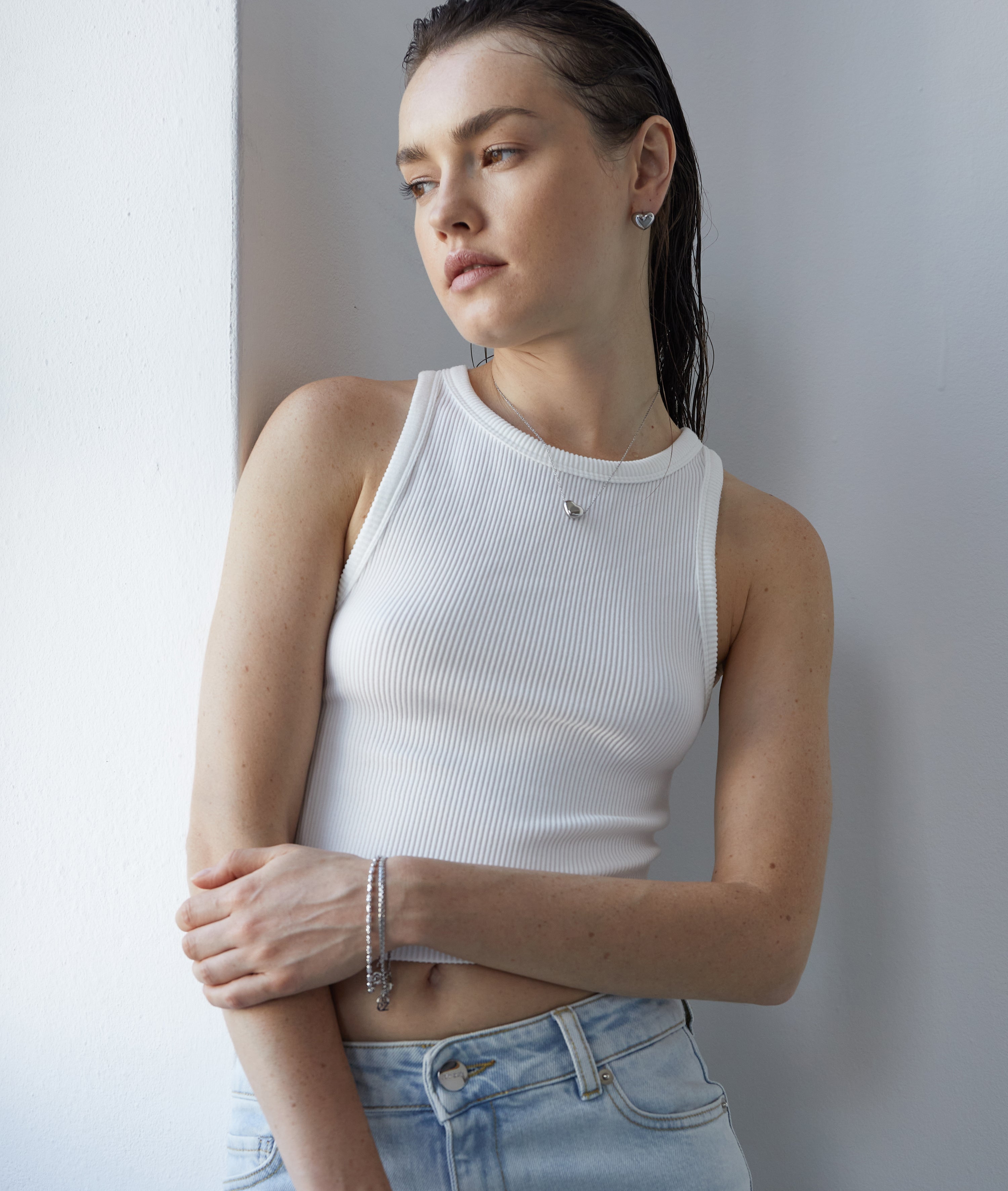 Eine Frau in einem weißen Tanktop und hellen Jeans steht an einer weißen Wand und blickt nachdenklich zur Seite. Sie trägt die Echtzeit Halskette - Puffy Heart von Echtzeit, die ihrem Look einen dezenten, zeitlosen Glanz verleiht.