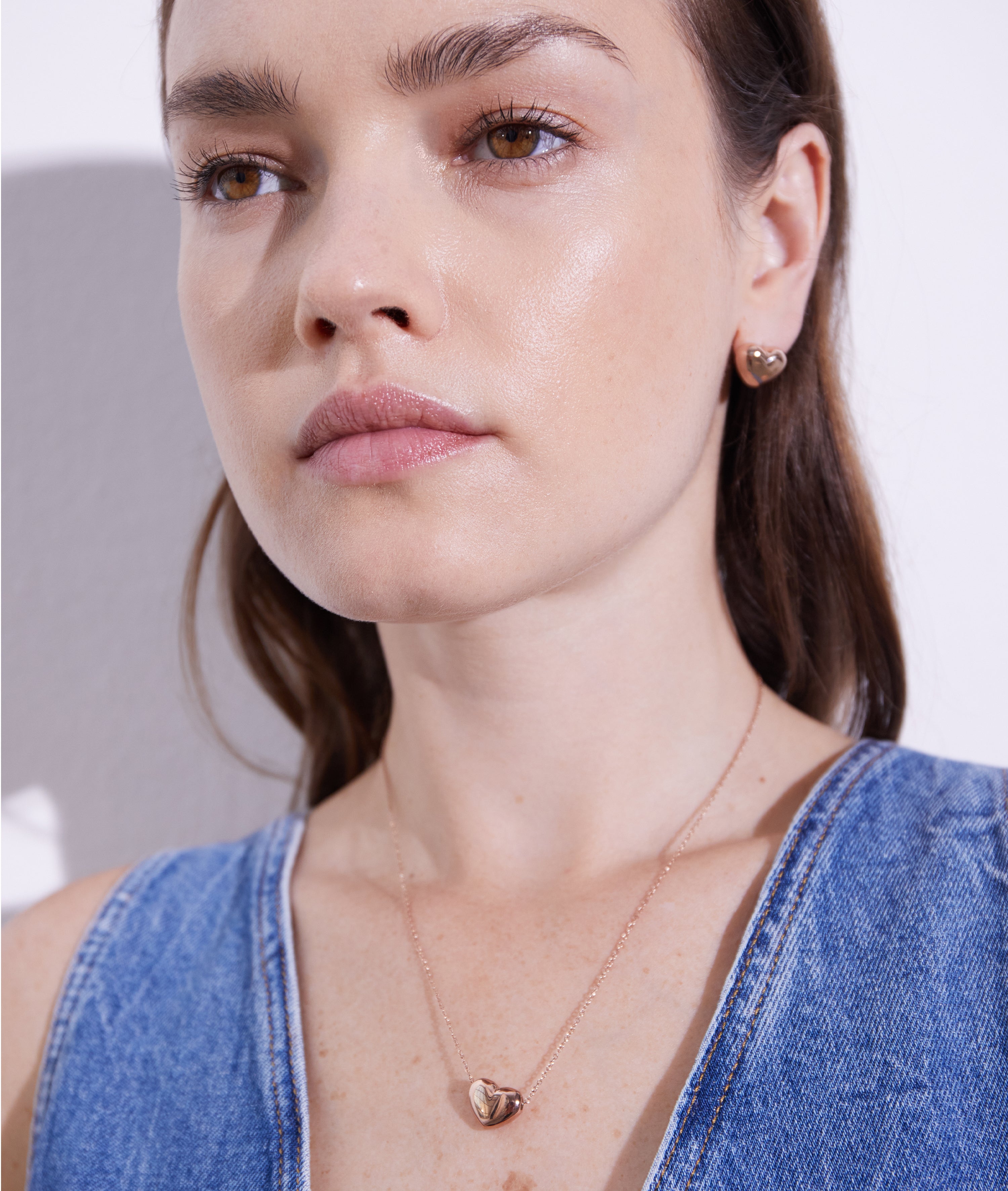 Eine Frau, die den Puffy Heart Echtzeit Ohrstecker und eine goldene Herzkette in einem blauen Denim-Top trägt, setzt einen romantischen Akzent vor einem weißen Hintergrund.