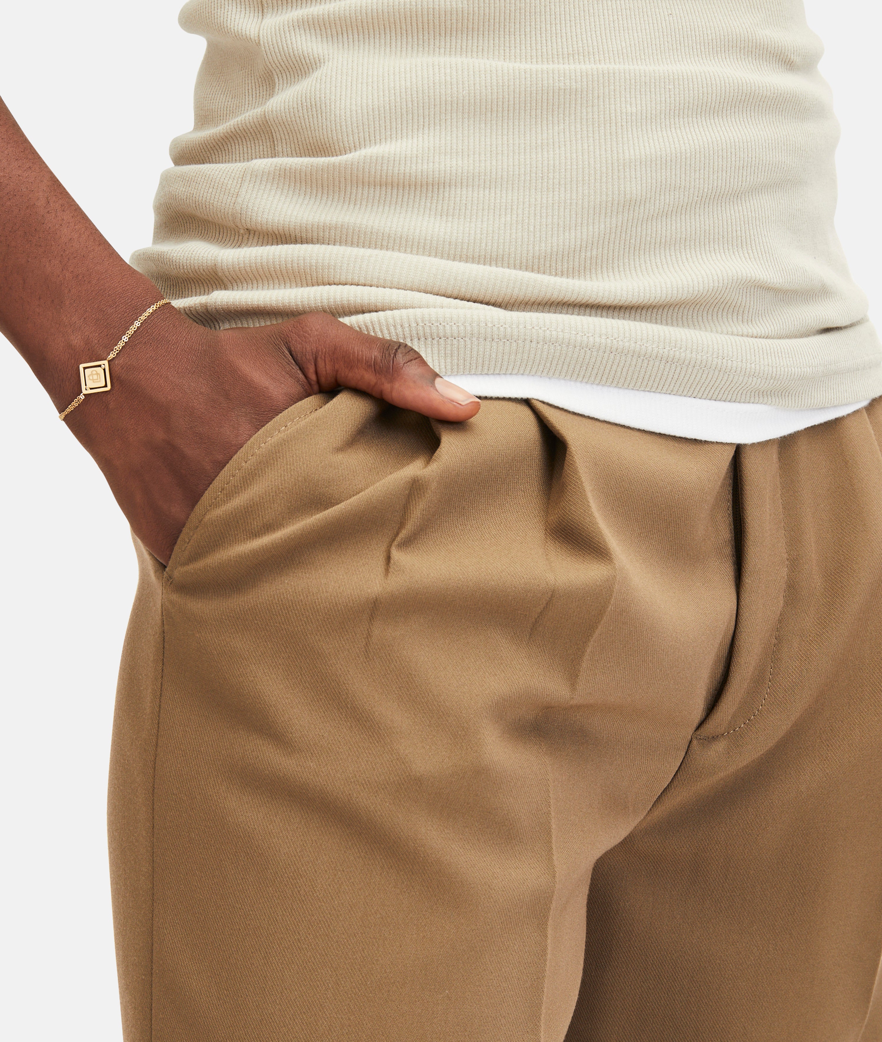 Eine Person, die eine hellbraune Hose und ein cremefarbenes, geripptes Oberteil trägt, mit einer Hand in der Tasche, ausgestattet mit dem goldenen LIEBESKIND BERLIN Armband - Swinging Icon von Liebeskind Berlin.