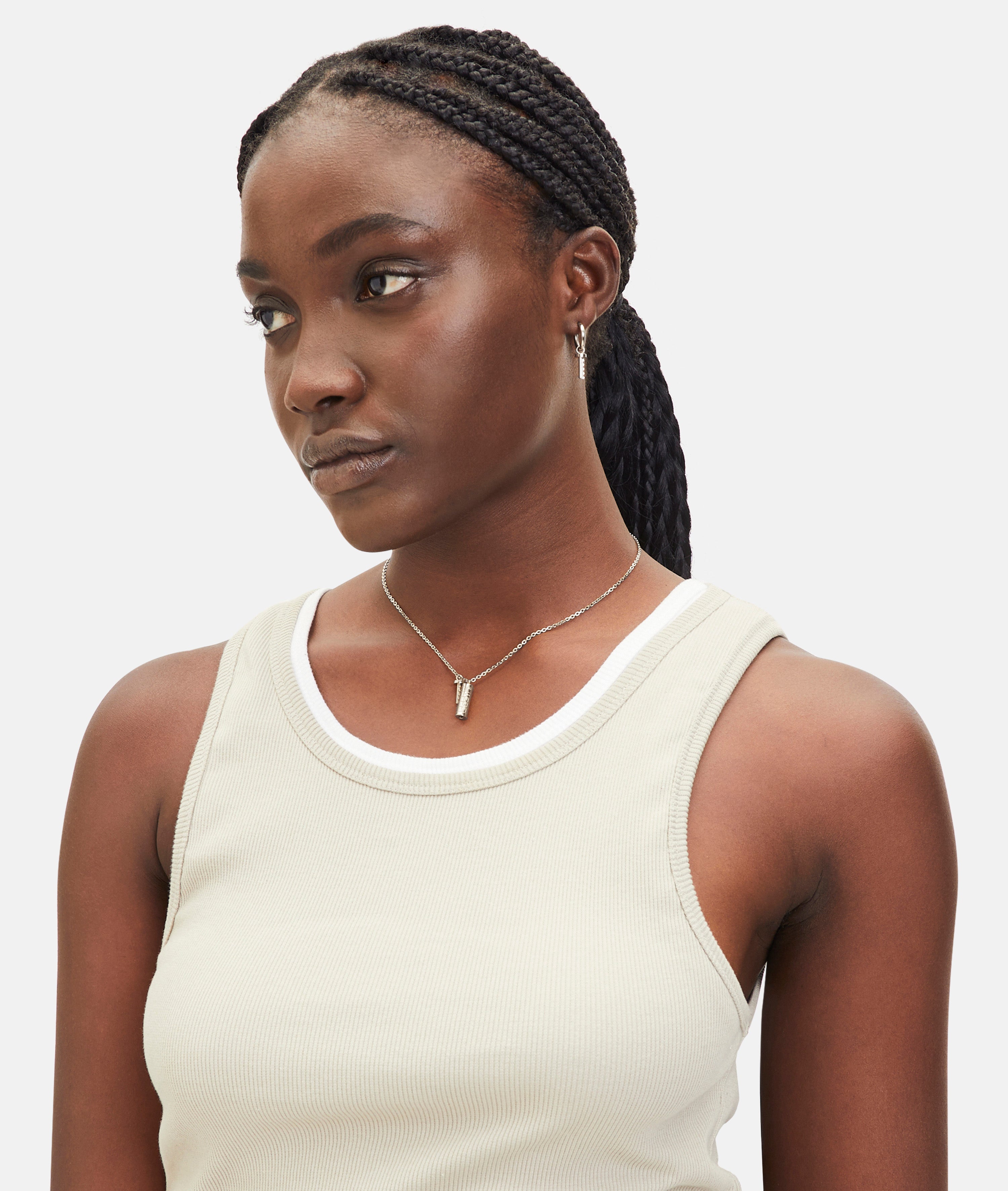 Eine Frau in einem beigen Tank-Top mit geflochtenen Haaren schaut zur Seite und trägt die LIEBESKIND BERLIN Halskette - The Love Lock. Diese elegante Halskette aus Edelstahl von Liebeskind Berlin ist ein dezentes Accessoire, perfekt als Geschenk Liebe.