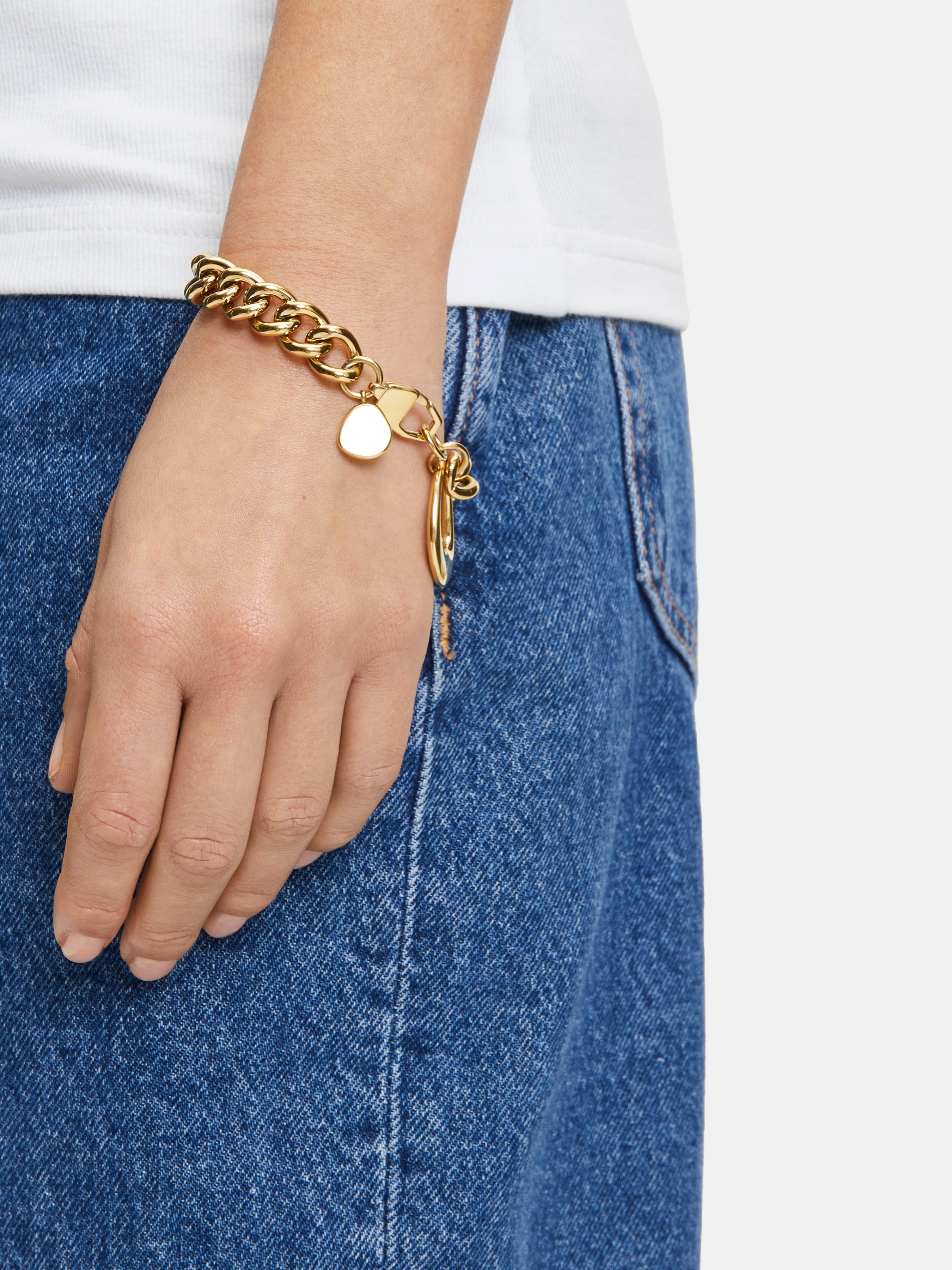 Eine Hand, die das Liebeskind Berlin Armband - The Organic, ein klobiges Edelstahlarmband mit einem Herzanhänger, trägt, ruht auf einer blauen Jeans und einem weißen Hemd.