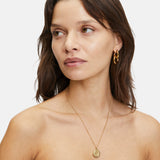 Die braunhaarige Frau trägt goldene Ohrringe, eine Liebeskind Berlin "The Organic Pendant" Halskette und ein schwarzes trägerloses Top.
