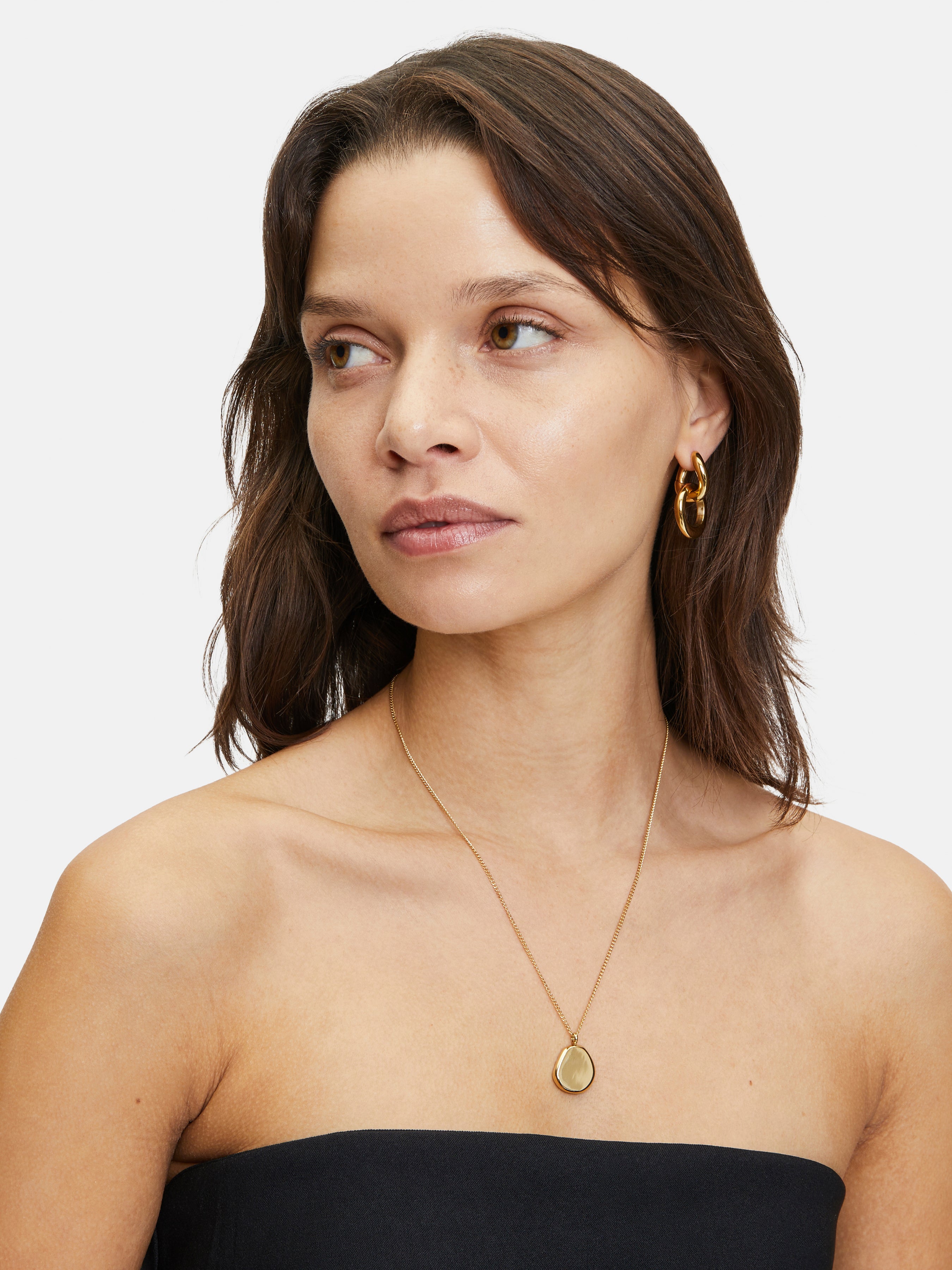 Die braunhaarige Frau trägt goldene Ohrringe, eine Liebeskind Berlin "The Organic Pendant" Halskette und ein schwarzes trägerloses Top.