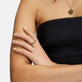 Frau in einem schwarzen trägerlosen Kleid mit verschränkten Armen, trägt einen goldenen Ring und die Liebeskind Berlin "The Organic Pendant" Halskette.