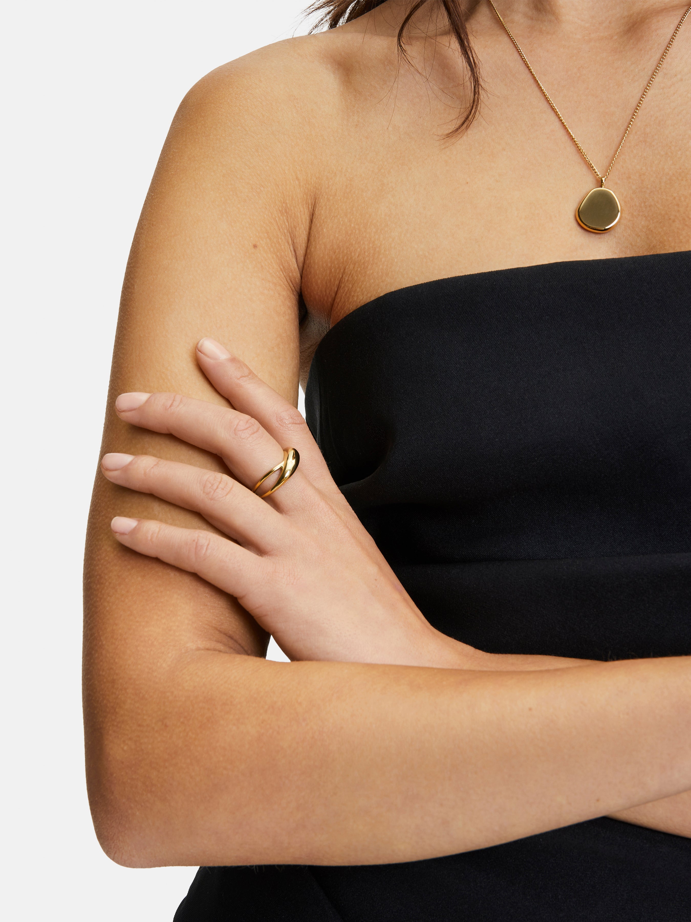 Frau in einem schwarzen trägerlosen Kleid mit verschränkten Armen, trägt einen goldenen Ring und die Liebeskind Berlin "The Organic Pendant" Halskette.