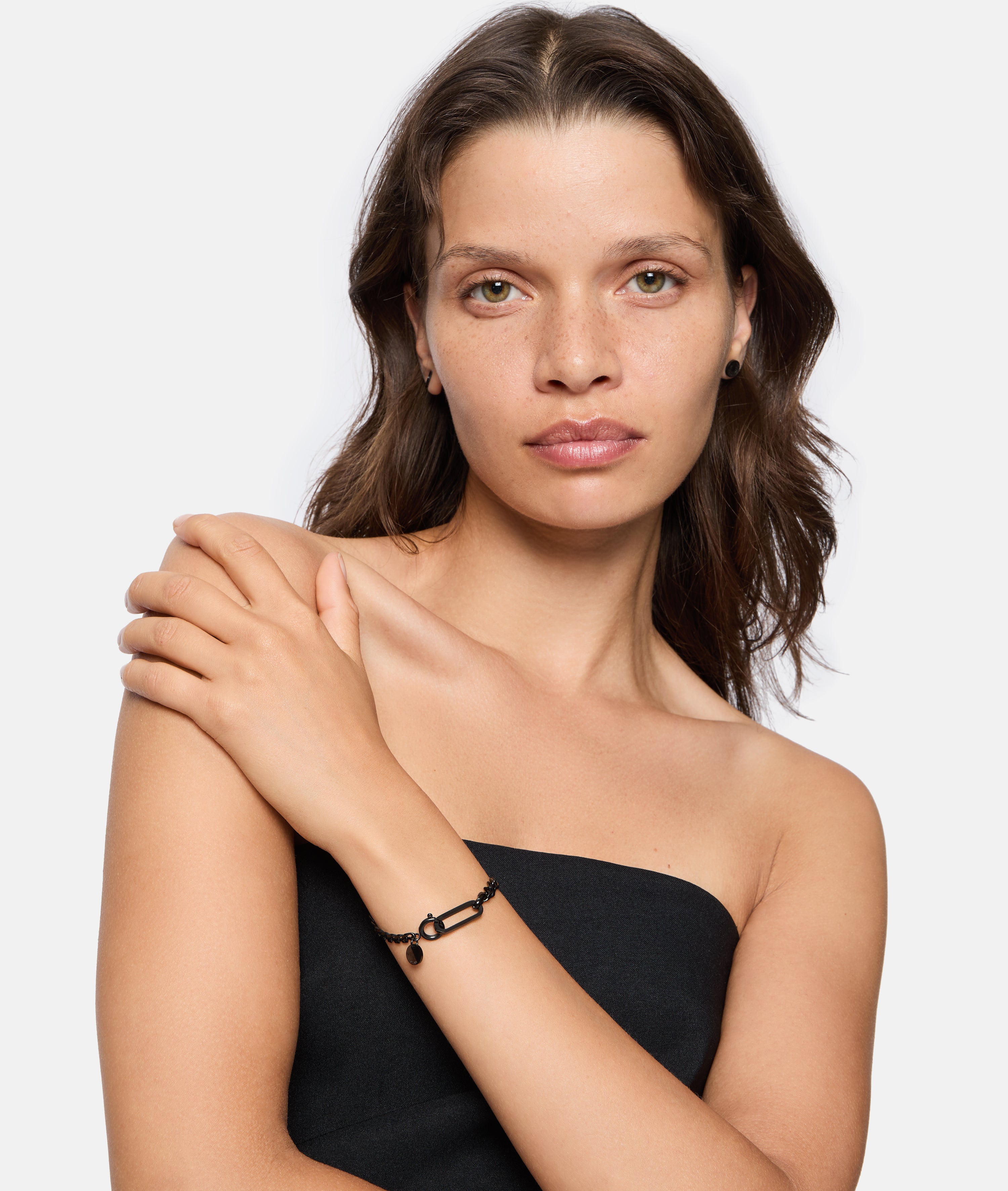 Frau mit gewellten braunen Haaren in einem trägerlosen schwarzen Oberteil posiert mit der Hand auf der Schulter und präsentiert mühelos den LIEBESKIND BERLIN Armband - Karabiner von Liebeskind Berlin, das Must-have Trend Accessoire Schmuck dieser Saison.