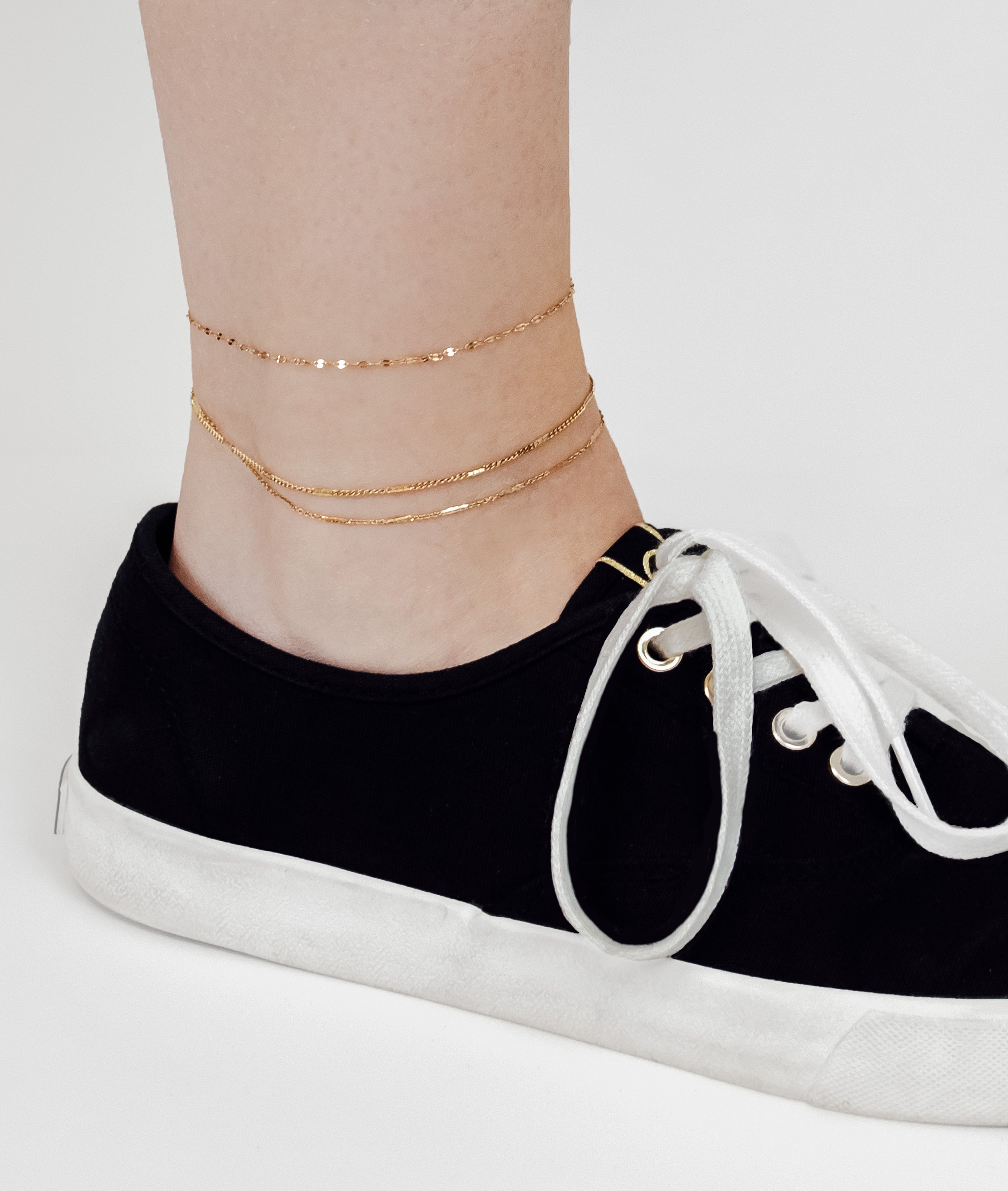 Eine Person trägt schwarze Turnschuhe und die LIEBESKIND BERLIN Fußkette - Sparkling With Layers von Liebeskind Berlin als schickes Accessoire an ihrem nackten Knöchel vor einem weißen Hintergrund.