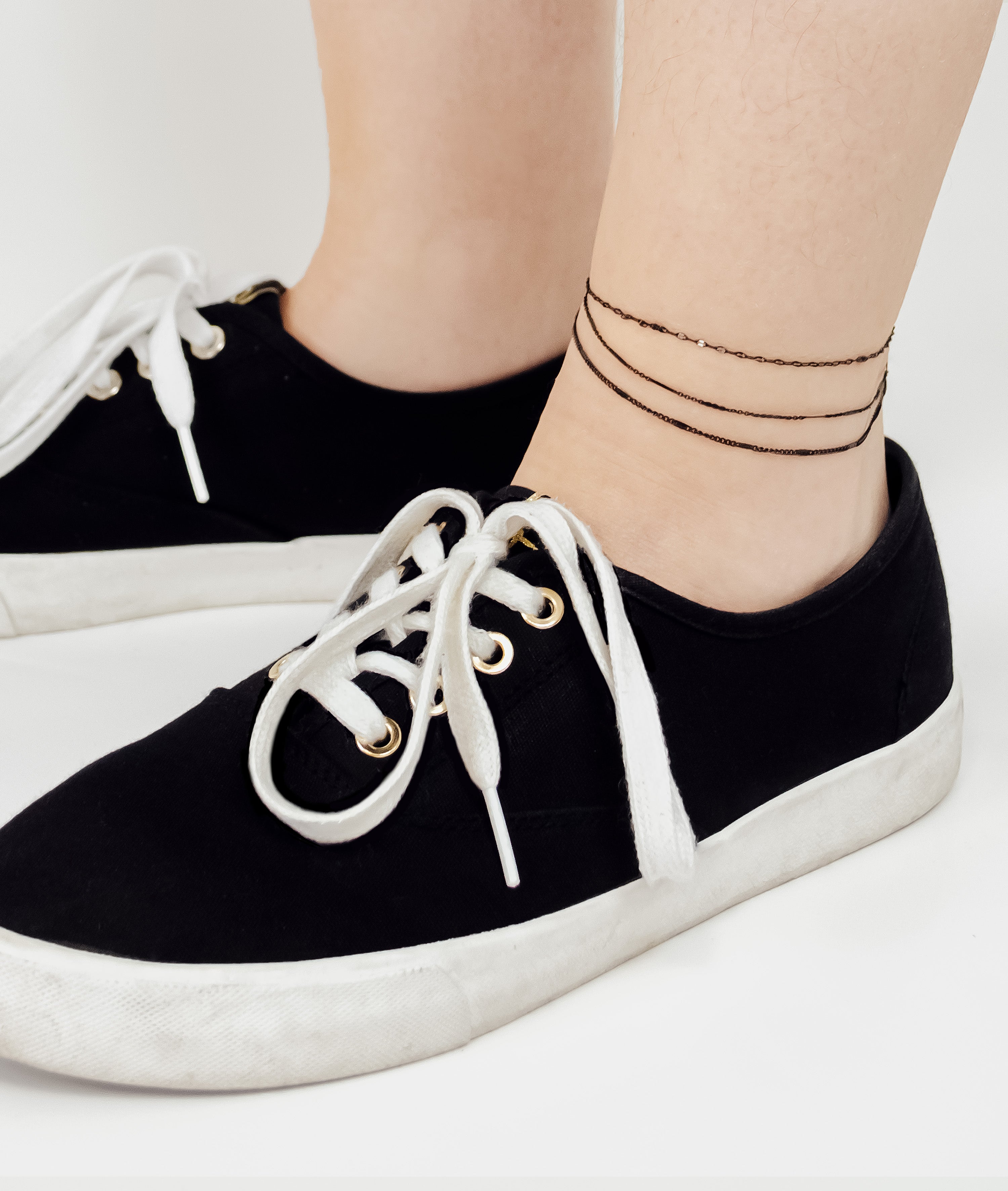 Nahaufnahme von Füßen in schwarzen Turnschuhen und der LIEBESKIND BERLIN Fußkette - Sparkling With Layers von Liebeskind Berlin, einem eleganten, mehrschichtigen Fußkettchen, vor hellem Hintergrund.