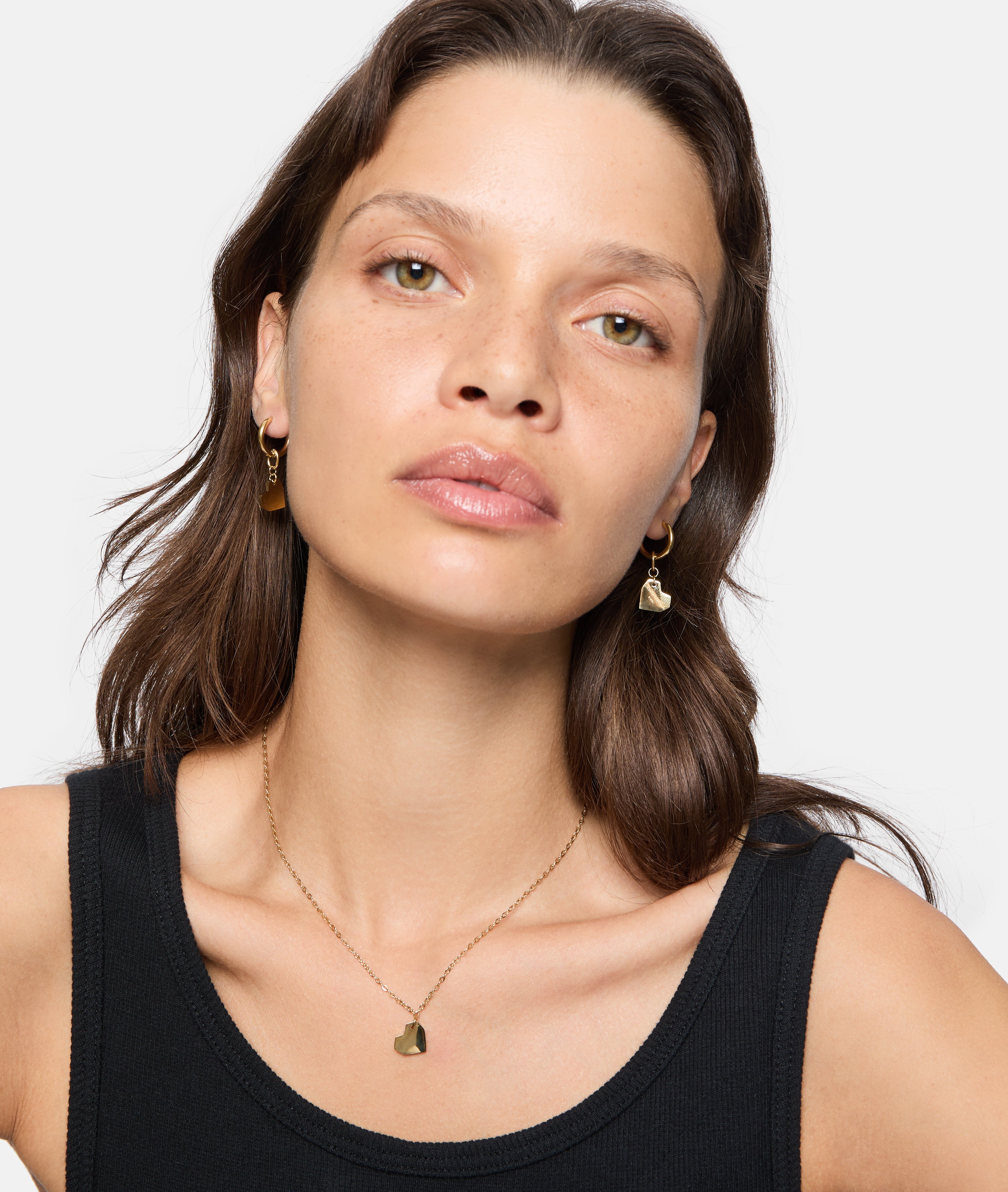 Eine Frau mit gewelltem braunem Haar, goldenen Ohrringen in Herzform und der LIEBESKIND BERLIN Halskette - Urban Heart von Liebeskind Berlin in einem schwarzen Tank-Top - eine Verkörperung von zeitloser Eleganz.