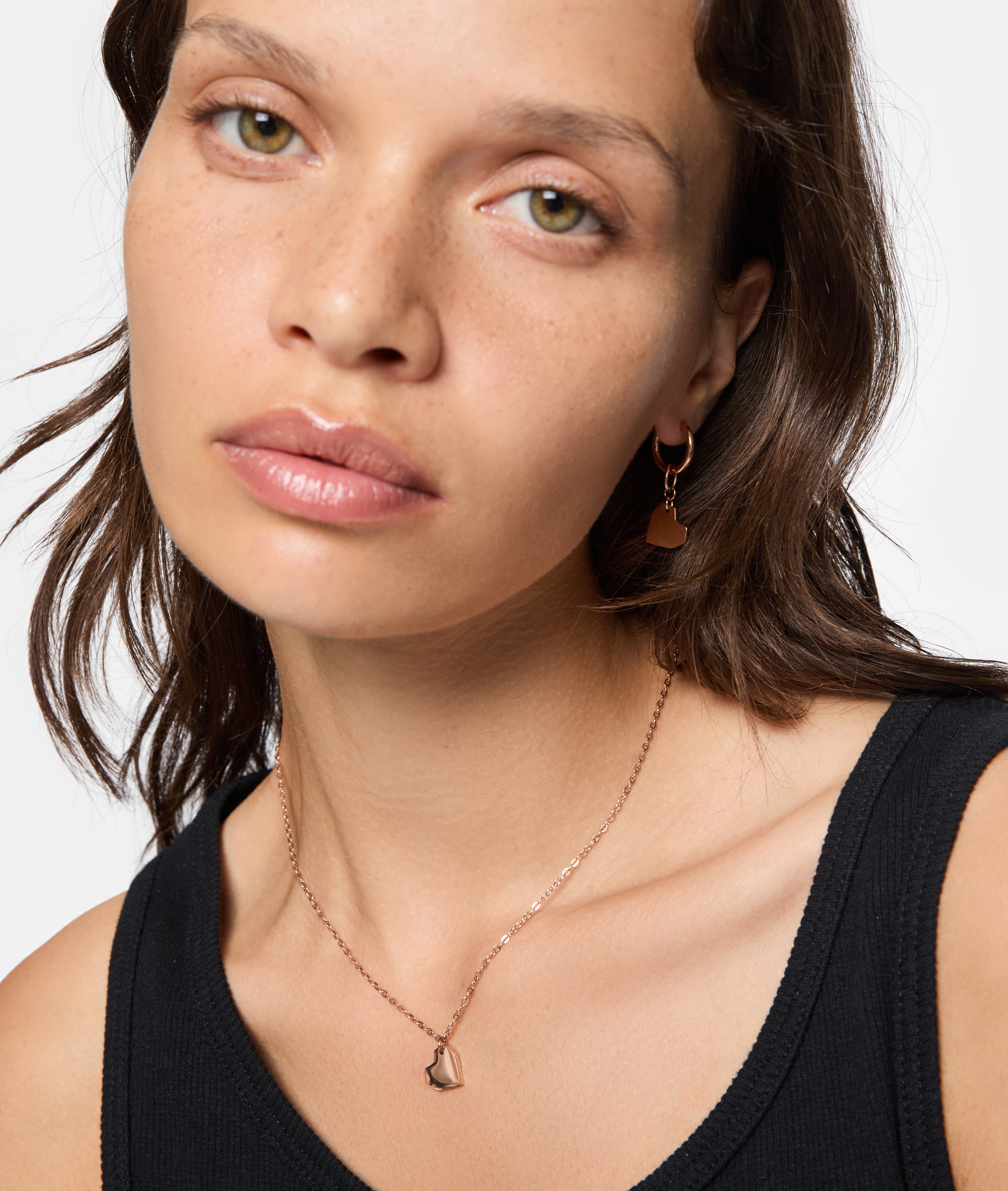 Frau mit schwarzem Oberteil, die Liebeskind Berlin Schmuck - eine goldene Halskette und LIEBESKIND BERLIN Creole - Urban Heart Ohrringe - trägt, blickt in die Kamera.