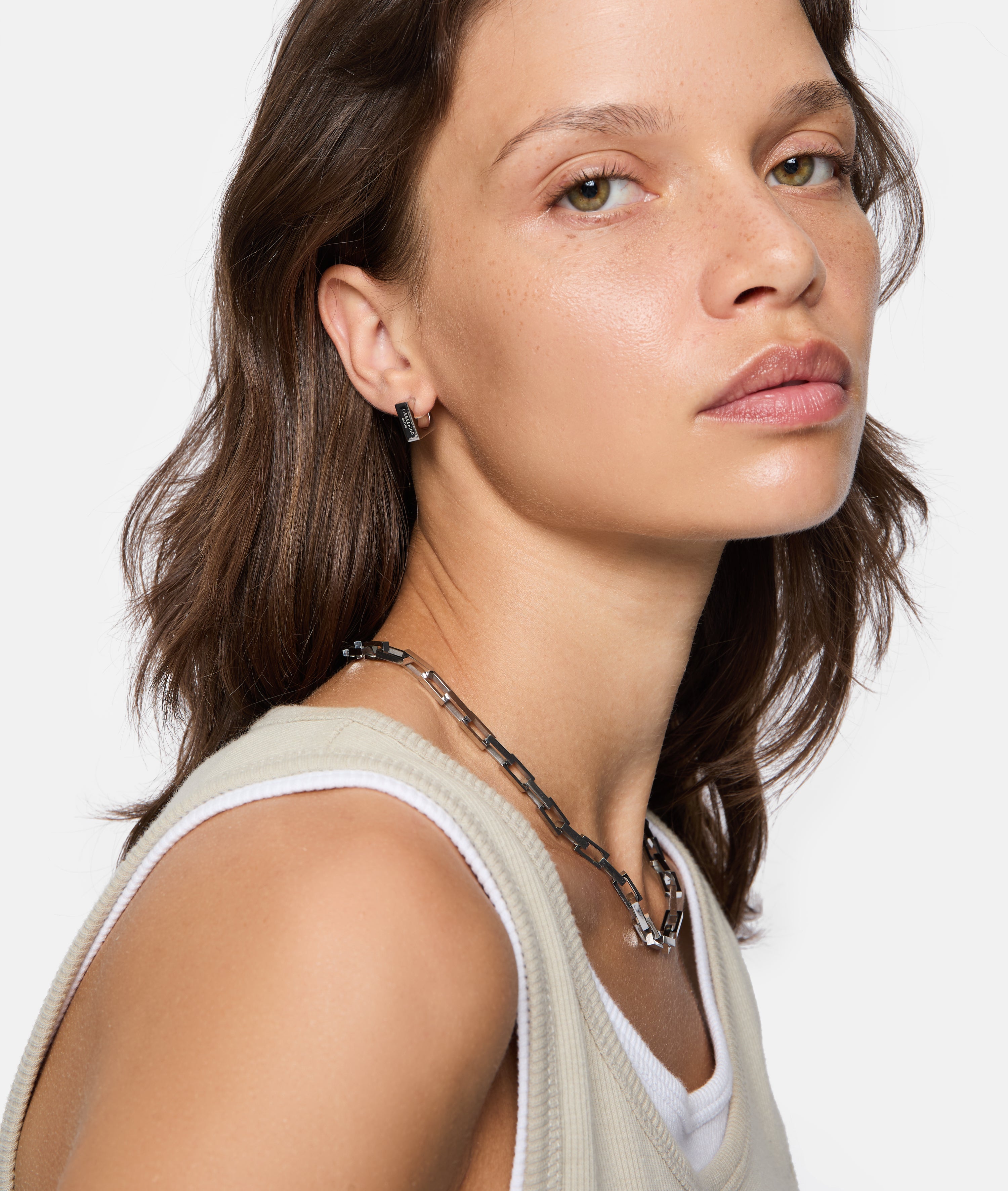 Frau mit braunen Haaren trägt ein ärmelloses beigefarbenes Oberteil, Kettenhalsband und LIEBESKIND BERLIN Creole - Urban Chic Ohrringe von Liebeskind Berlin, nach vorne schauend.
