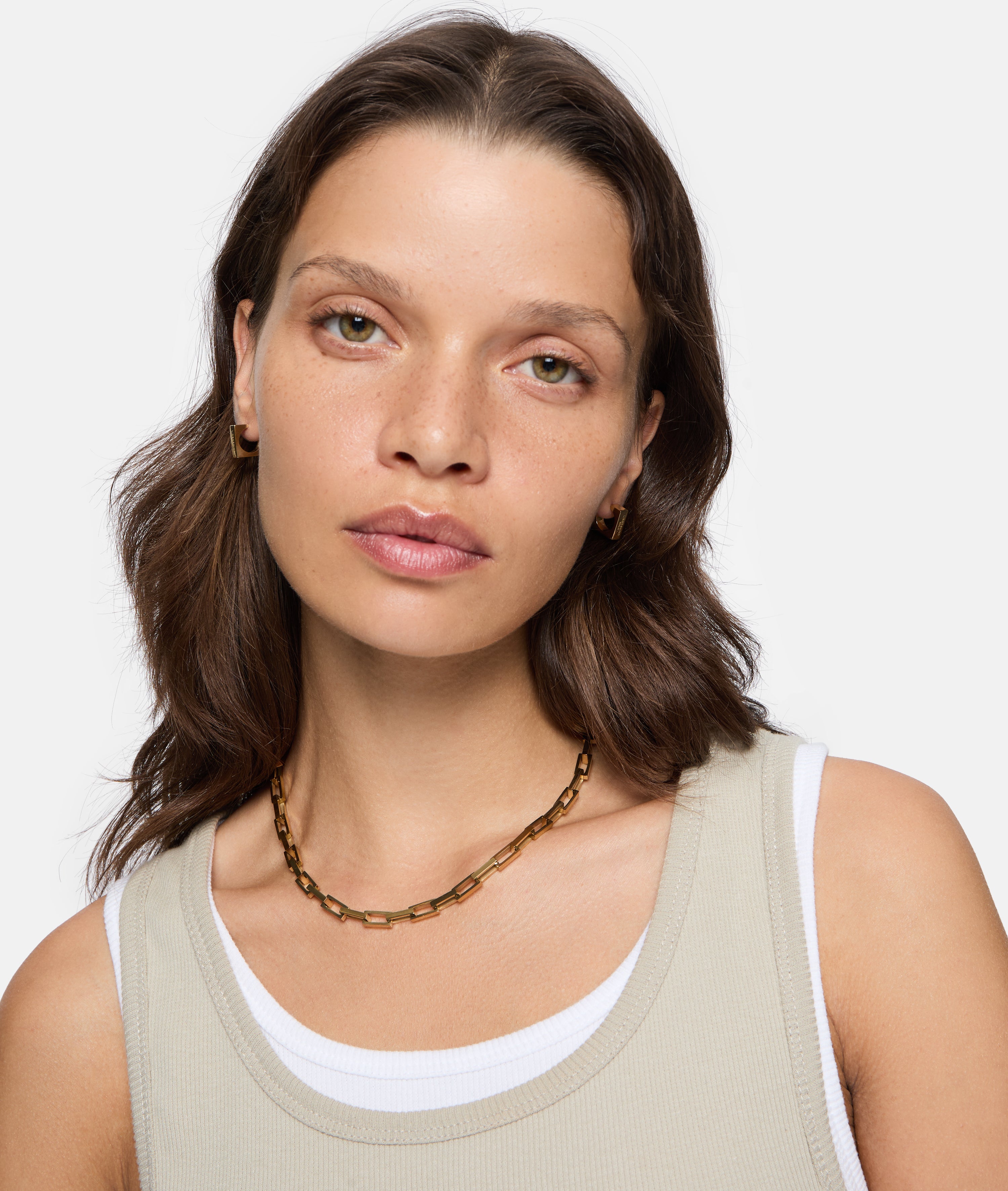 Frau mit gewellten braunen Haaren trägt LIEBESKIND BERLIN Creole - Urban Chic Ohrringe von Liebeskind Berlin, eine goldene Halskette und ein beigefarbenes Tanktop über einem weißen Hemd.